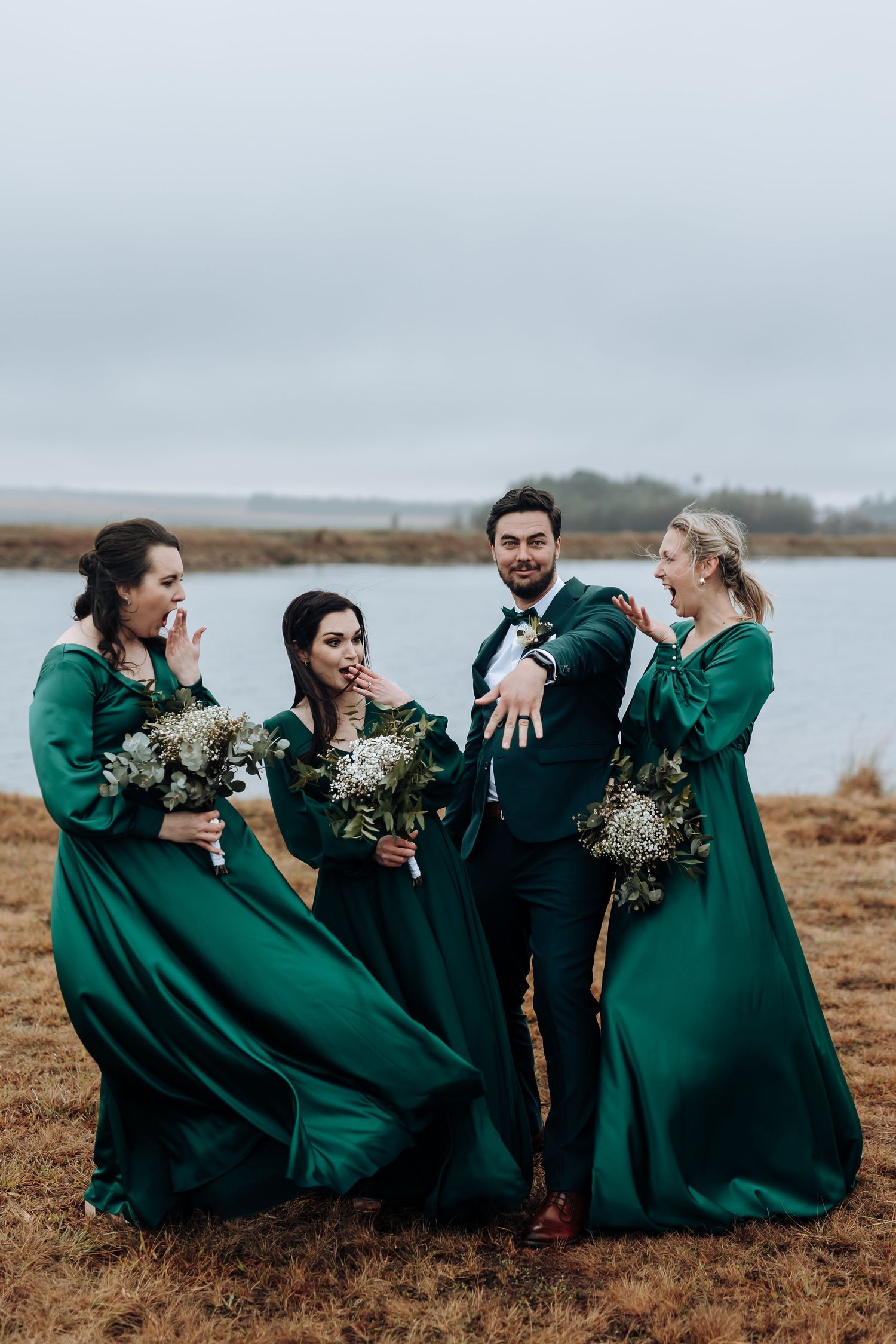 Wedding party in emerald attire share laughs by waterfront on cloudy day.