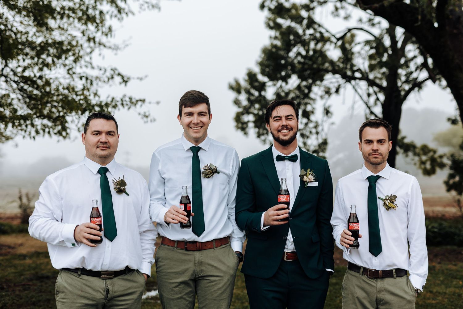 Groomsmen in coordinated outfits hold drinks while posing in a foggy outdoor setting.