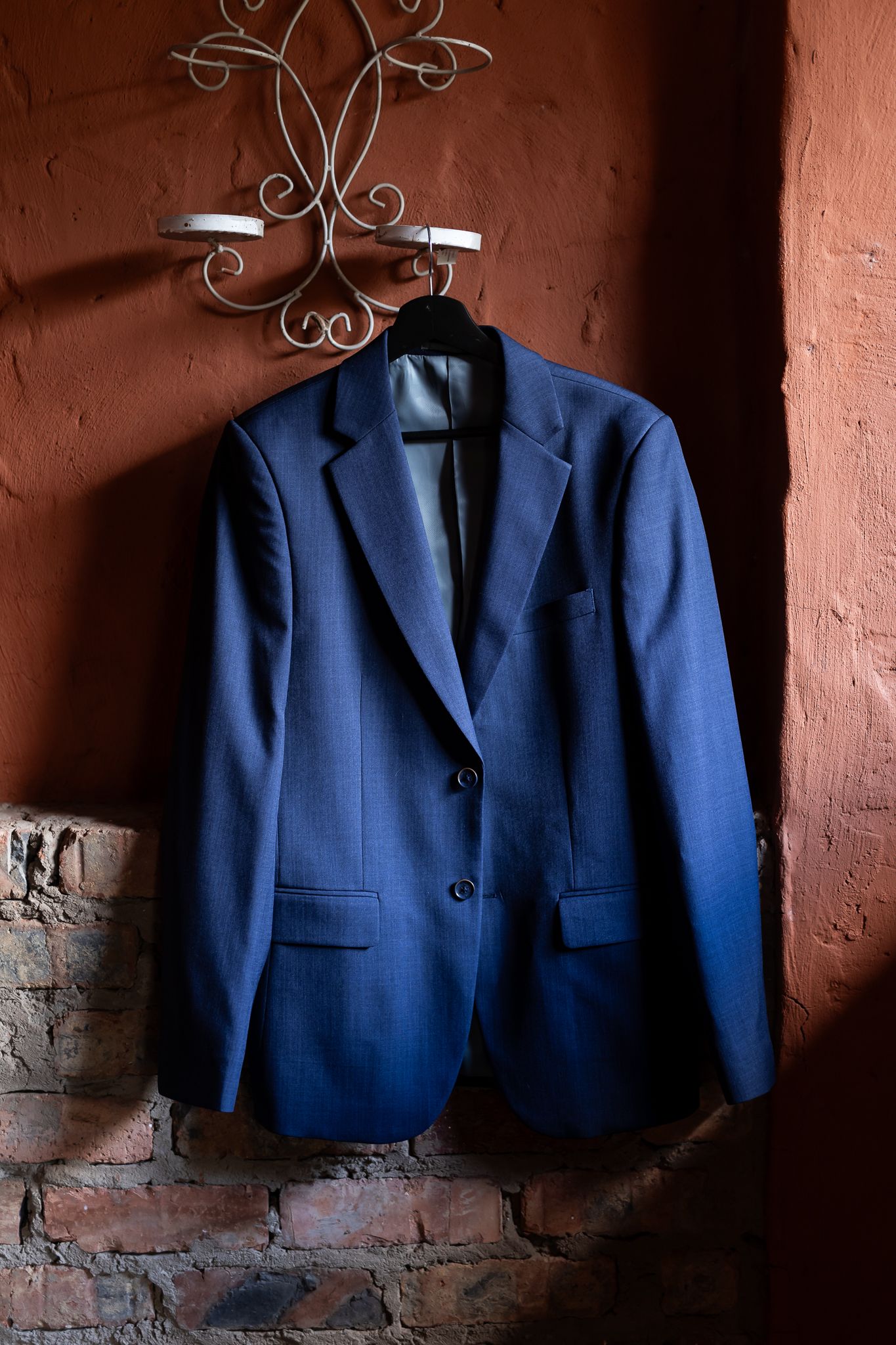 Navy blue suit jacket hanging on ornate metal wall hook against terracotta colored wall.