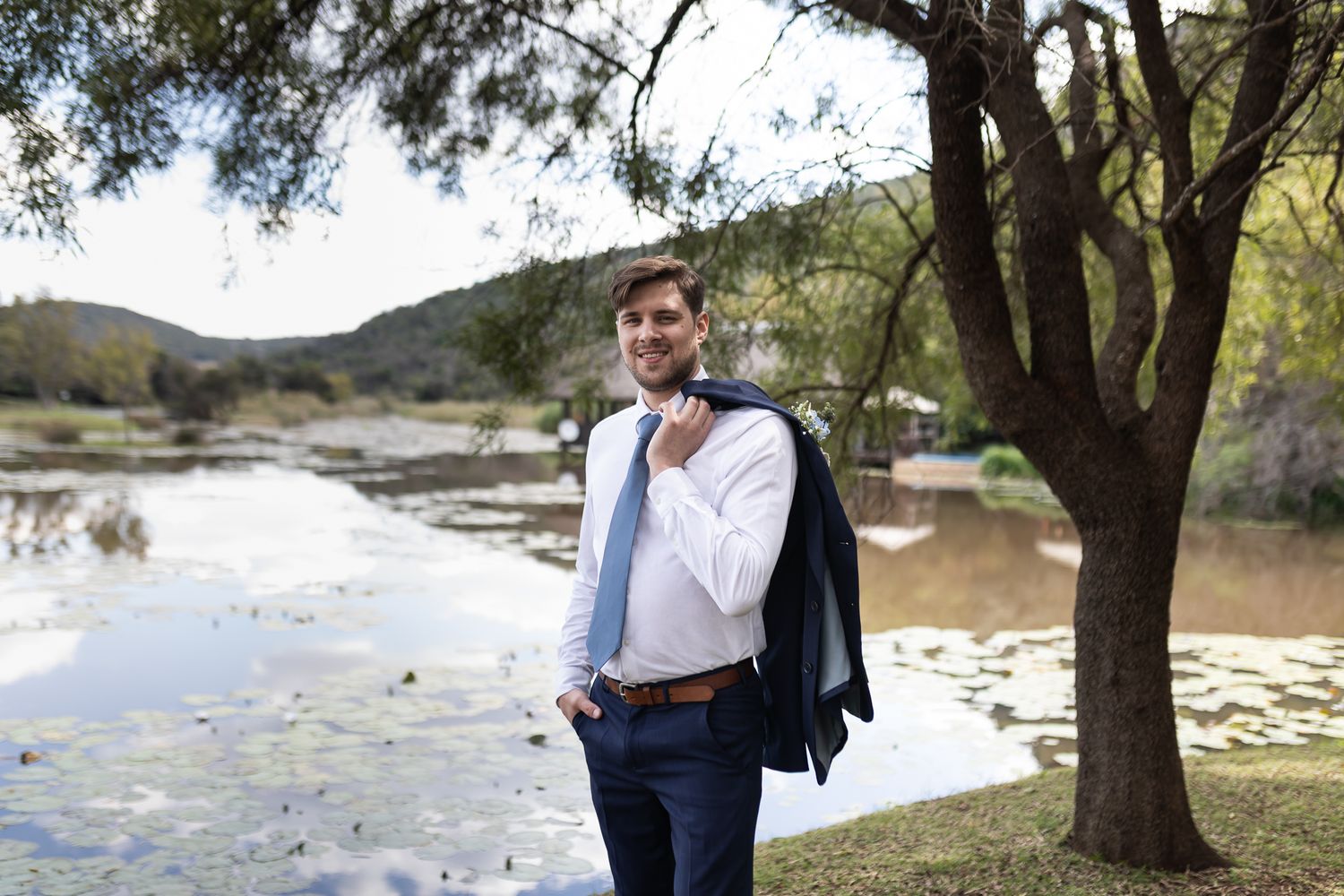 A groom in formal attire poses casually near a scenic lake with trees.