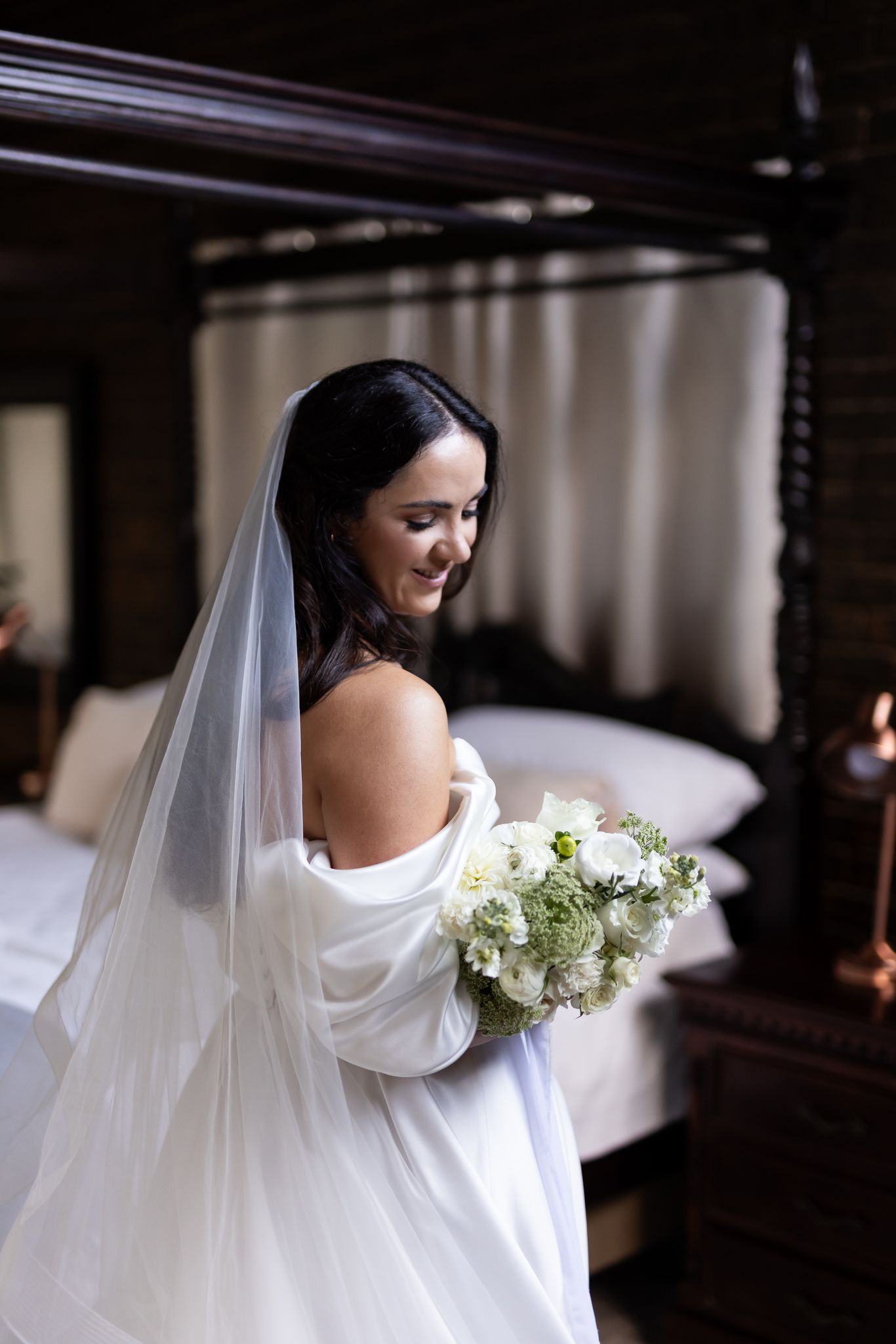 Bride in off-shoulder gown holds white bouquet while looking over shoulder.