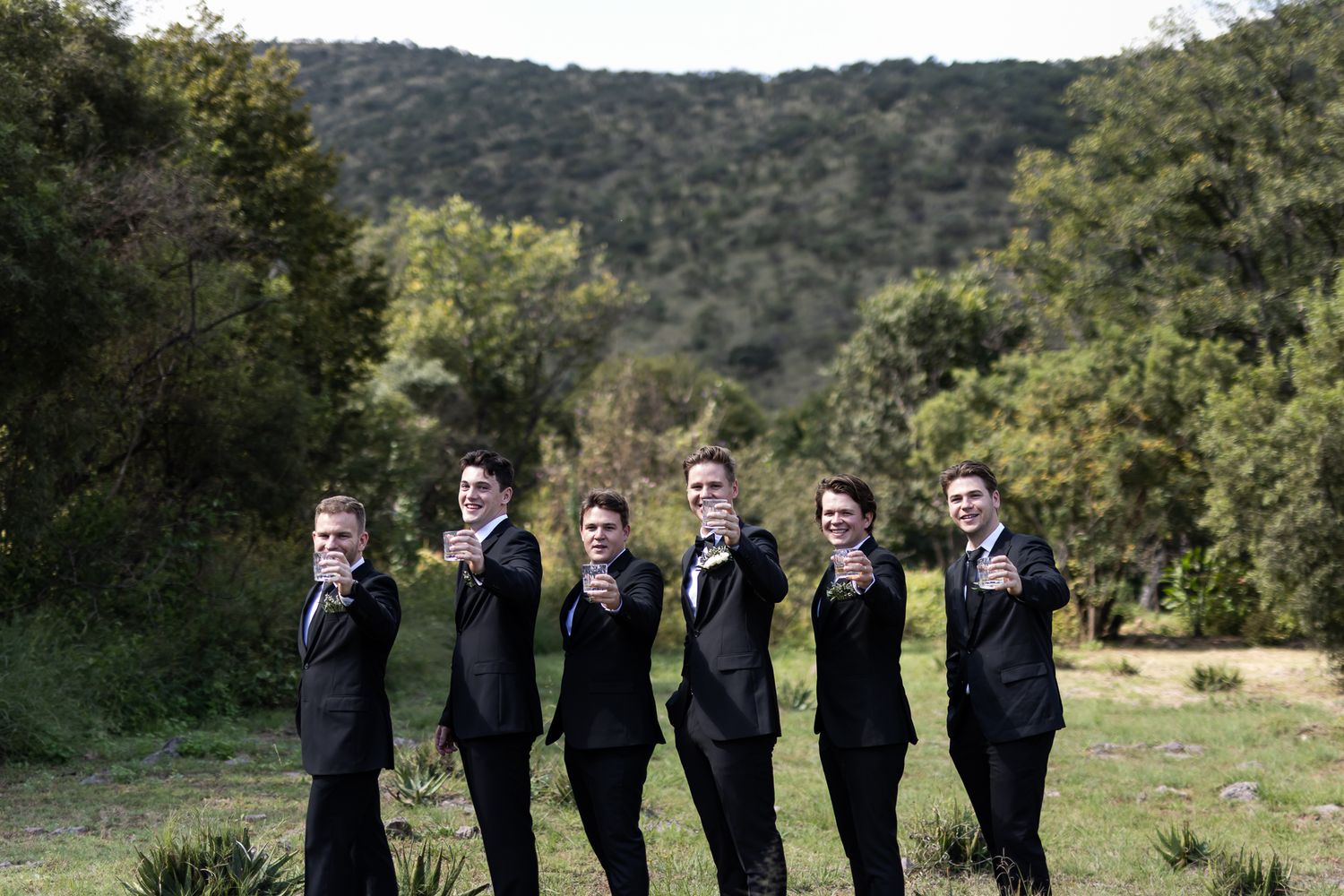 Wedding party in matching black suits lined up smoking cigars against a mountain landscape.