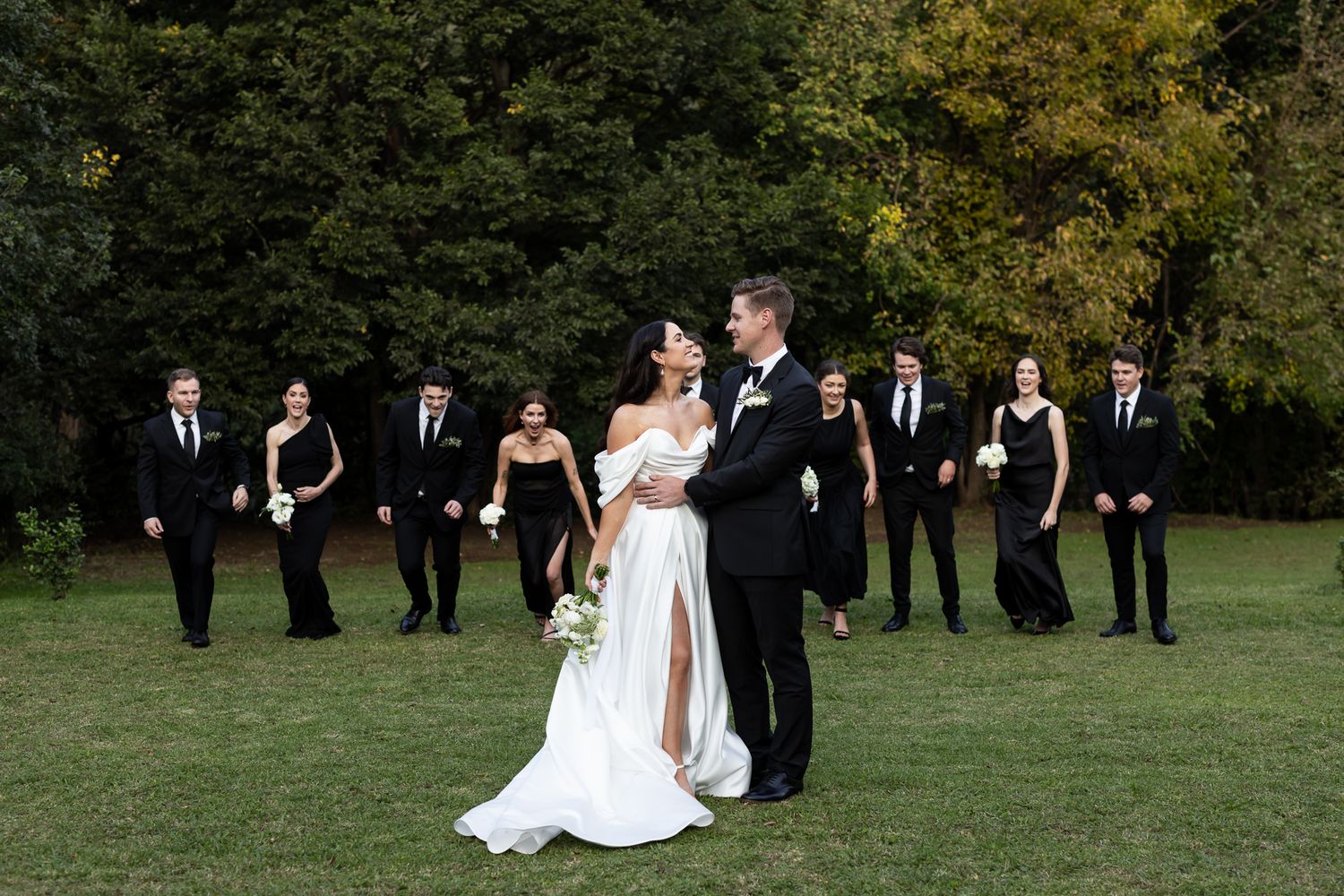 A wedding party in classic black attire poses elegantly on grass with fall foliage behind them.