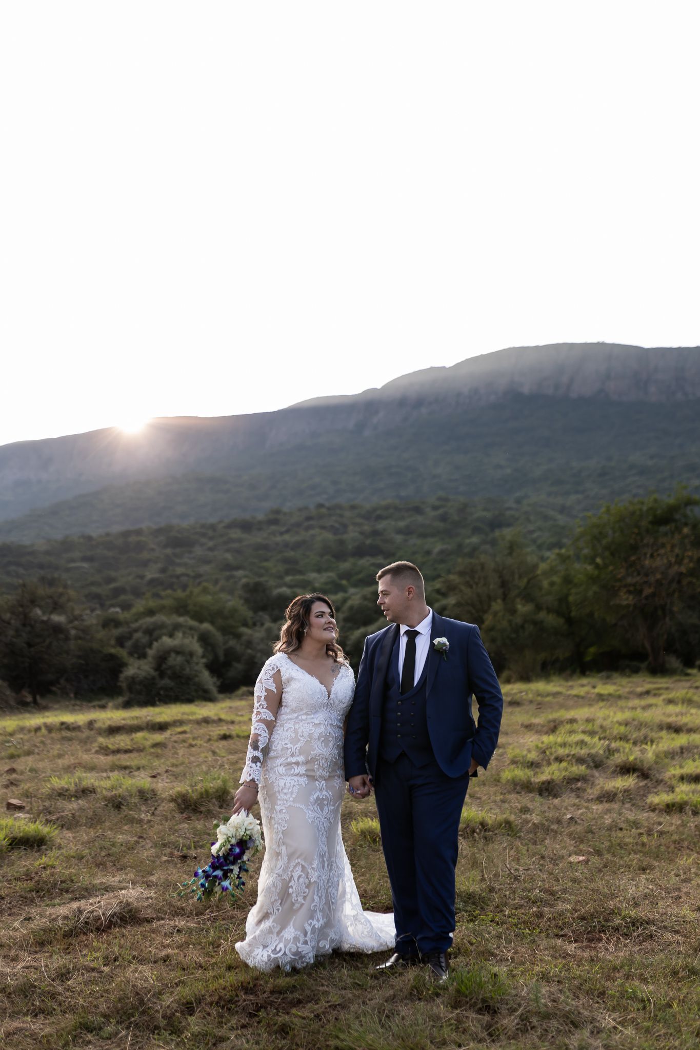 Wedding couple standing in mountain field at sunset holding colorful bouquet.