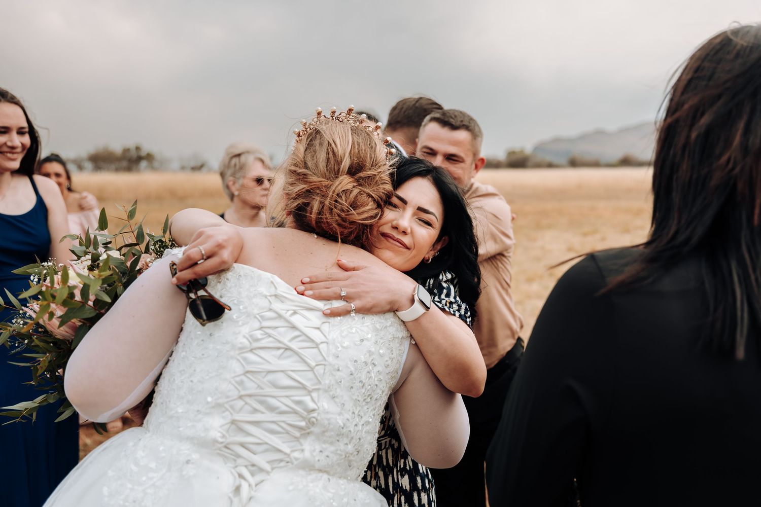 A group gathers outdoors sharing emotional hugs and embraces during a wedding celebration in a field.