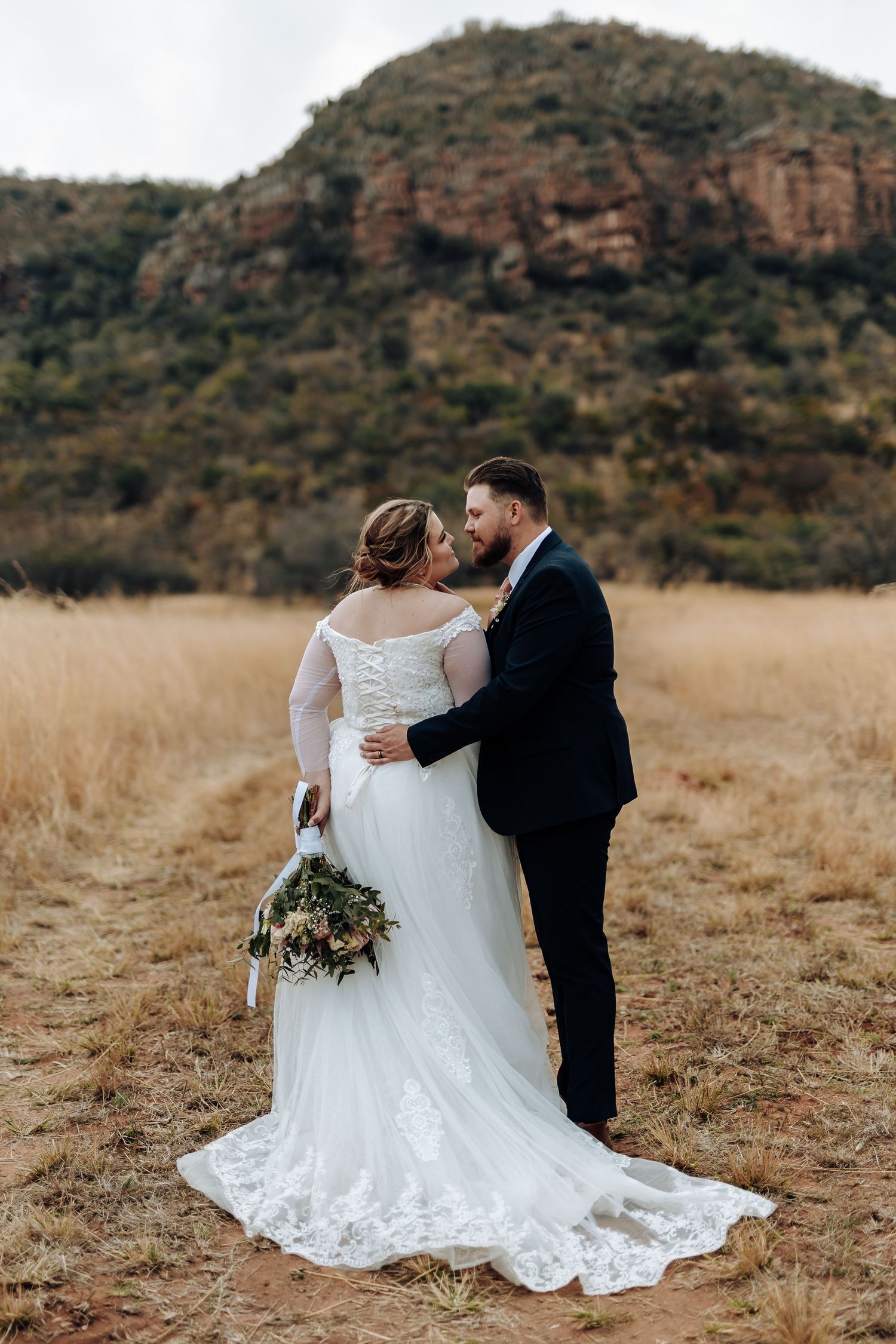 A couple embraces in rustic outdoor setting with mountain backdrop and tall wheat grass as sun sets.