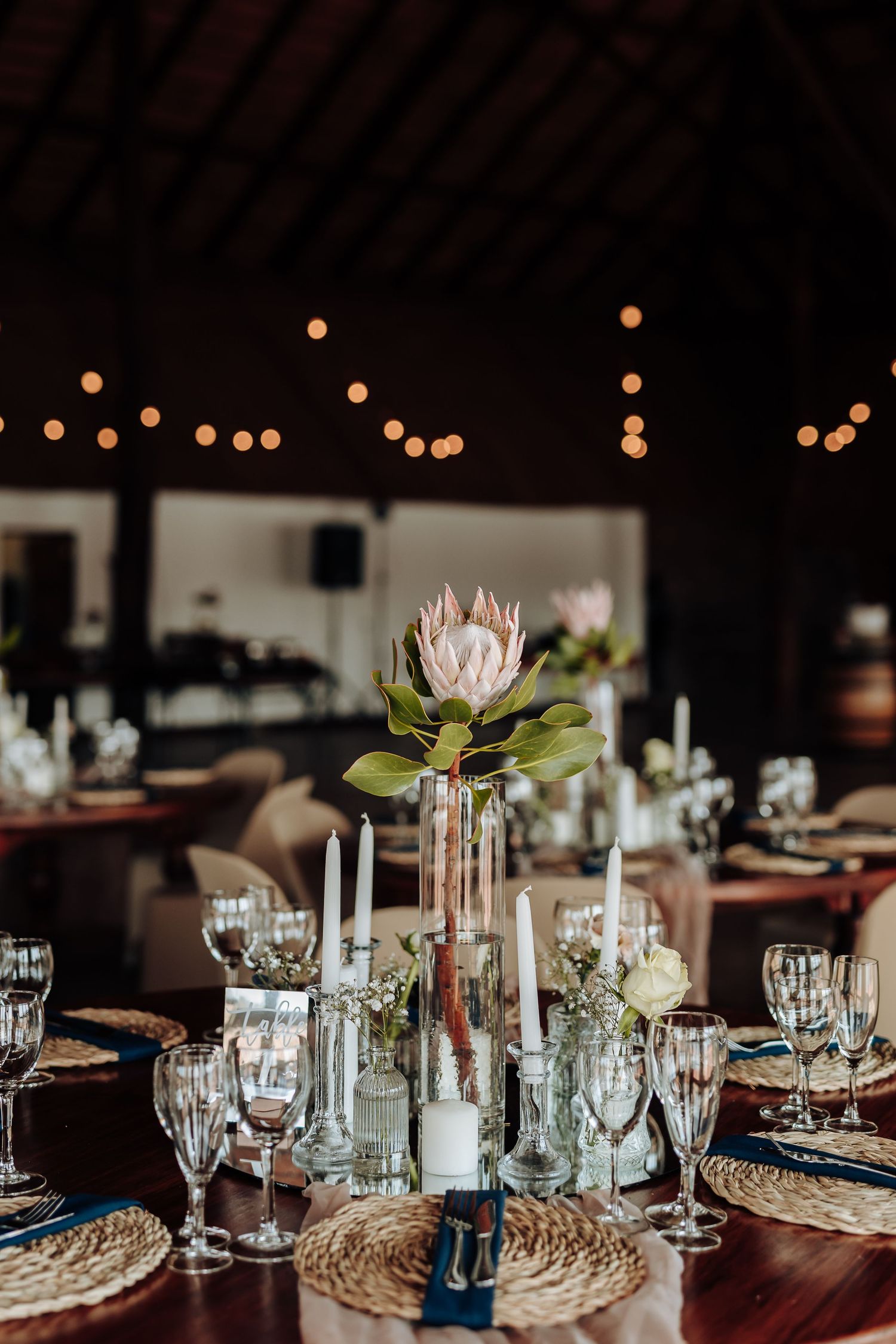 Elegant table setting with protea flower centerpiece, white candles, and woven placemats under string lights at reception.