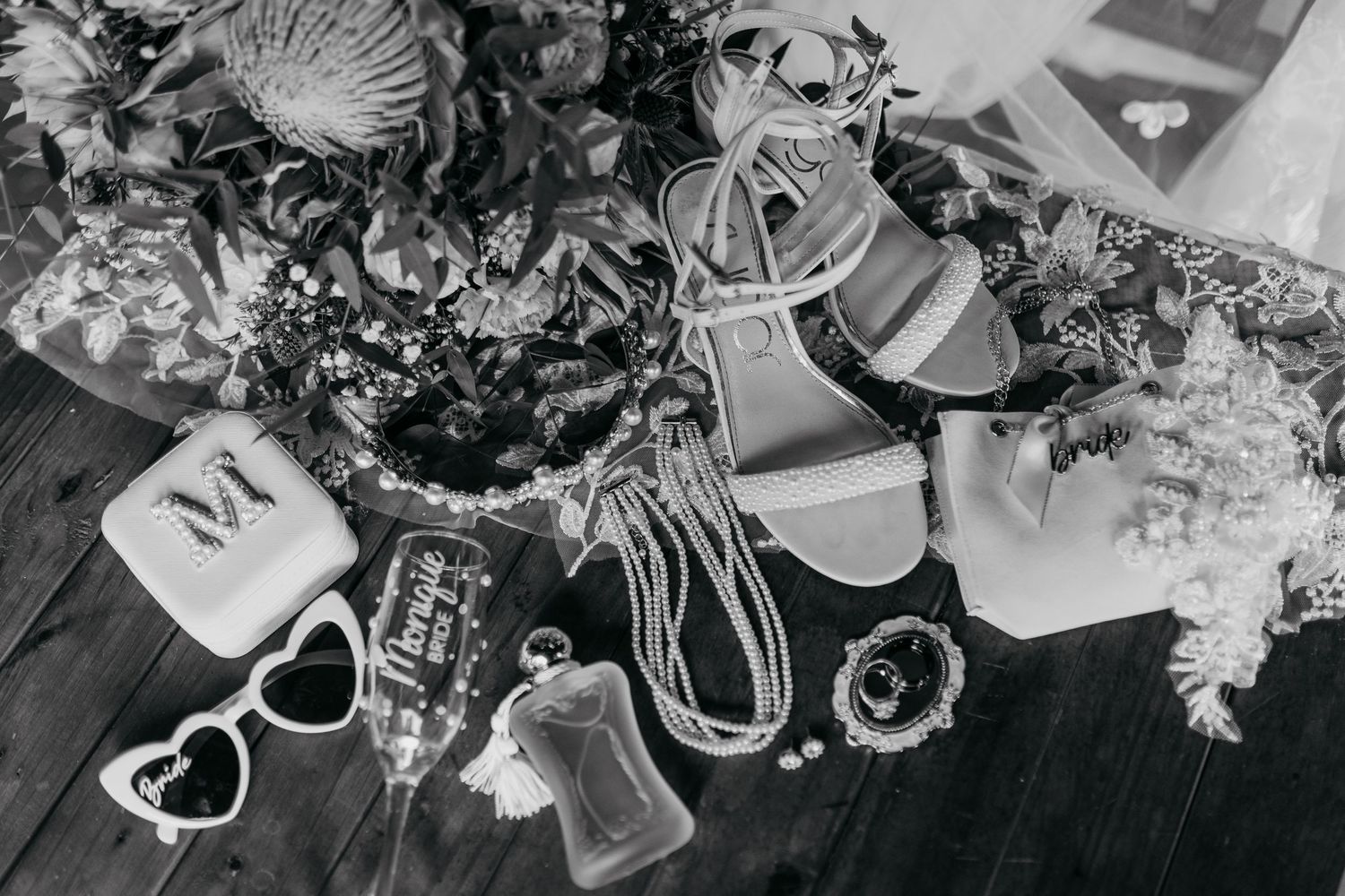 Black and white flat lay of wedding accessories including sandals, sunglasses, and floral arrangements on dark wood floor.