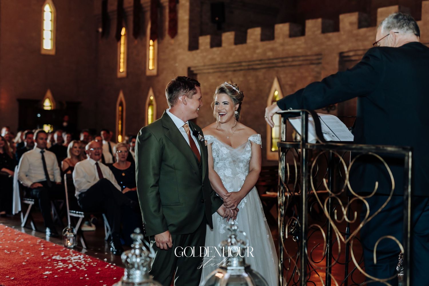 Bride in lace gown and groom in green suit exchange vows at a church altar with red carpet aisle.