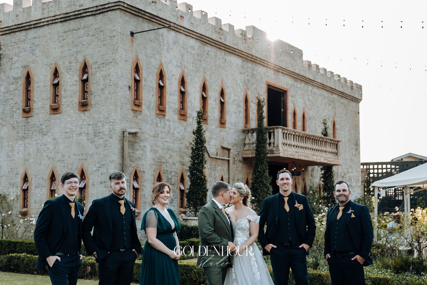 Wedding party posing in front of a castle-style venue at golden hour with bride and groom kissing.