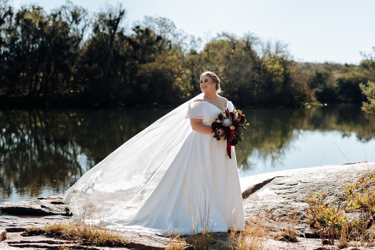 Bride in white off-shoulder ball gown holding bouquet by a reflective lake with trees in background.
