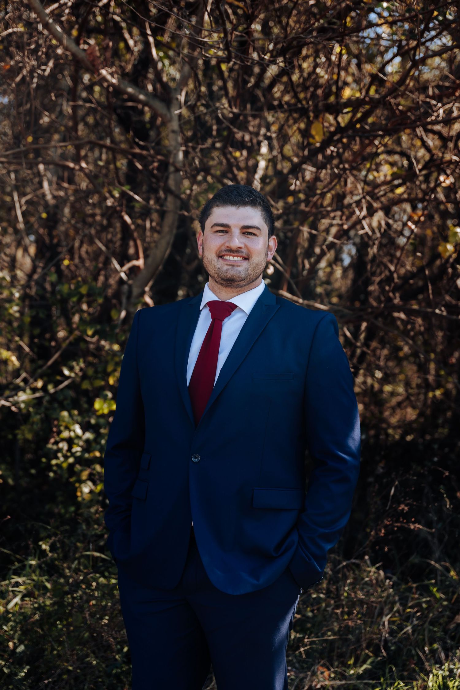 Man in navy blue suit and red tie smiling outdoors with autumn trees in background.