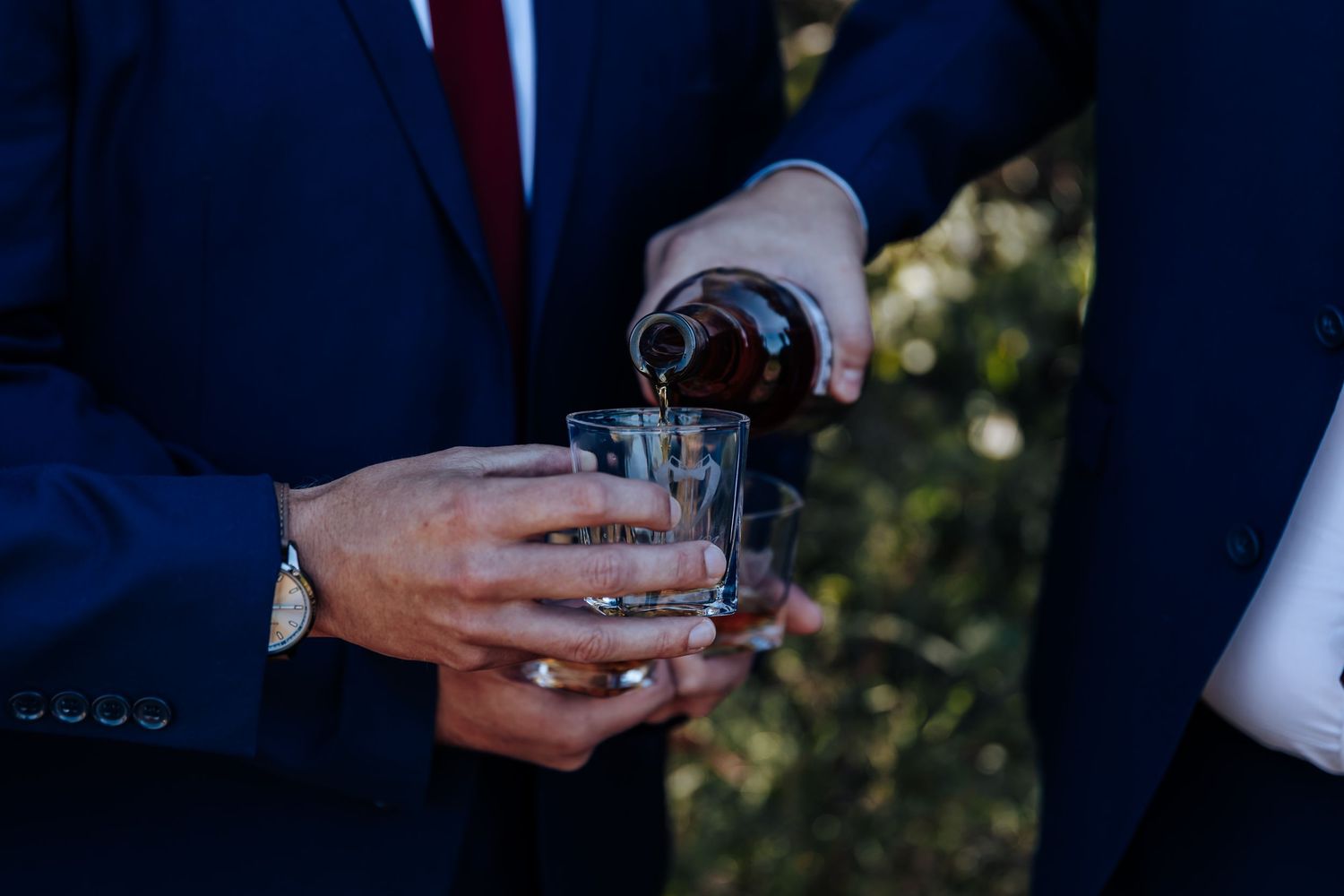 Man in blue suit pouring dark liquor from bottle into shot glasses at a formal event.