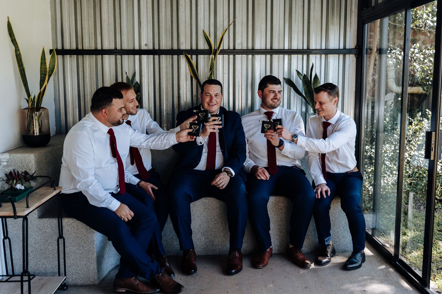 Five groomsmen in navy suits and red ties toasting with drinks at a wedding celebration.