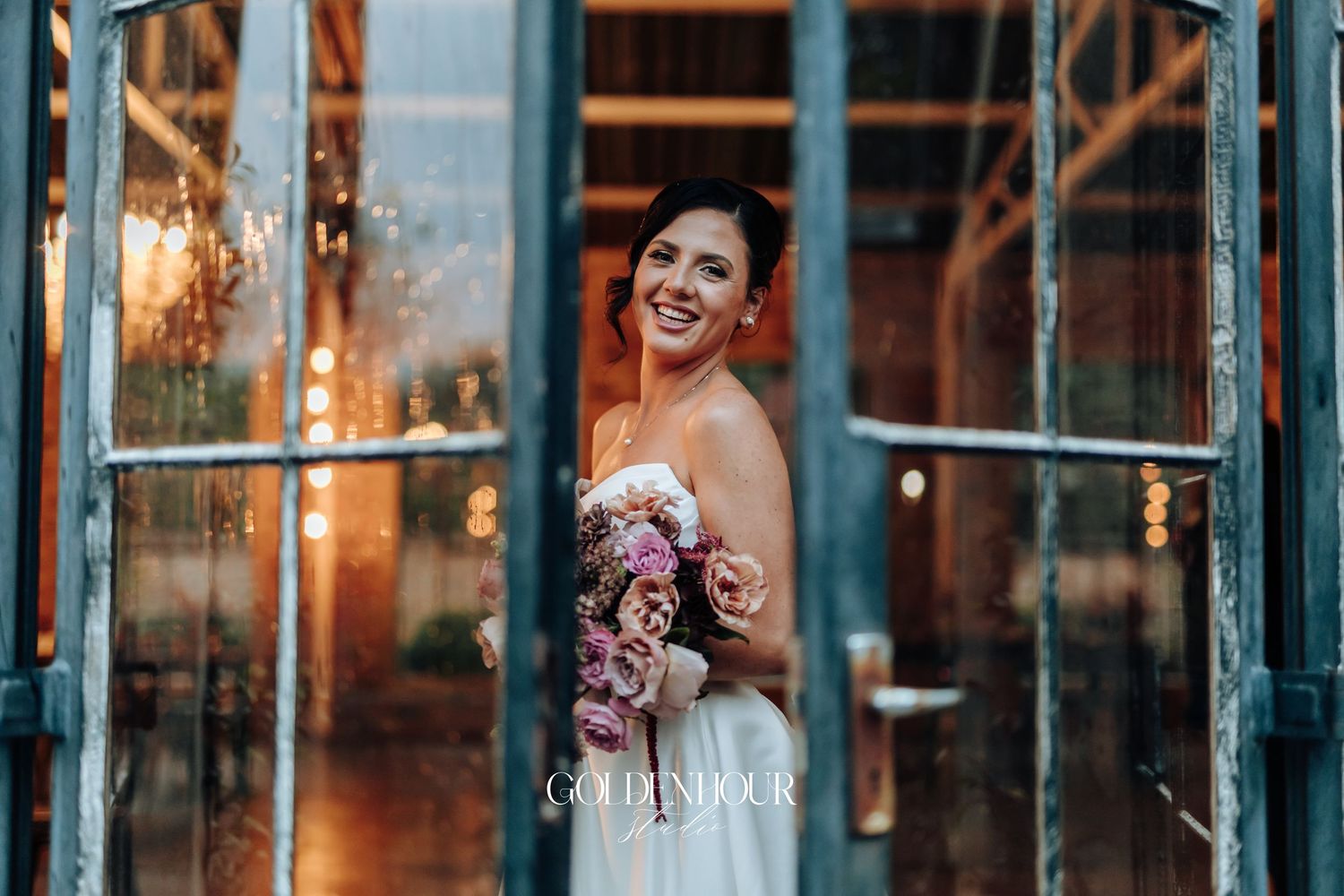 Glowing bride in strapless white gown smiling through glass doors holding a pink floral bouquet.