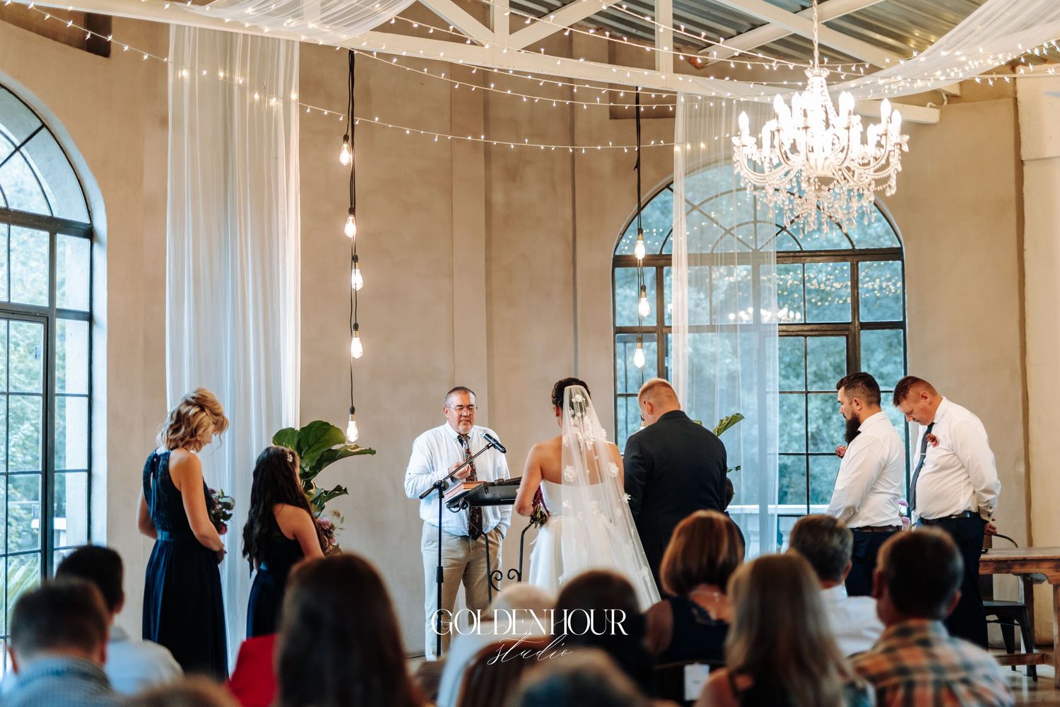 Wedding ceremony in elegant venue with chandelier, string lights, and live musician performing.