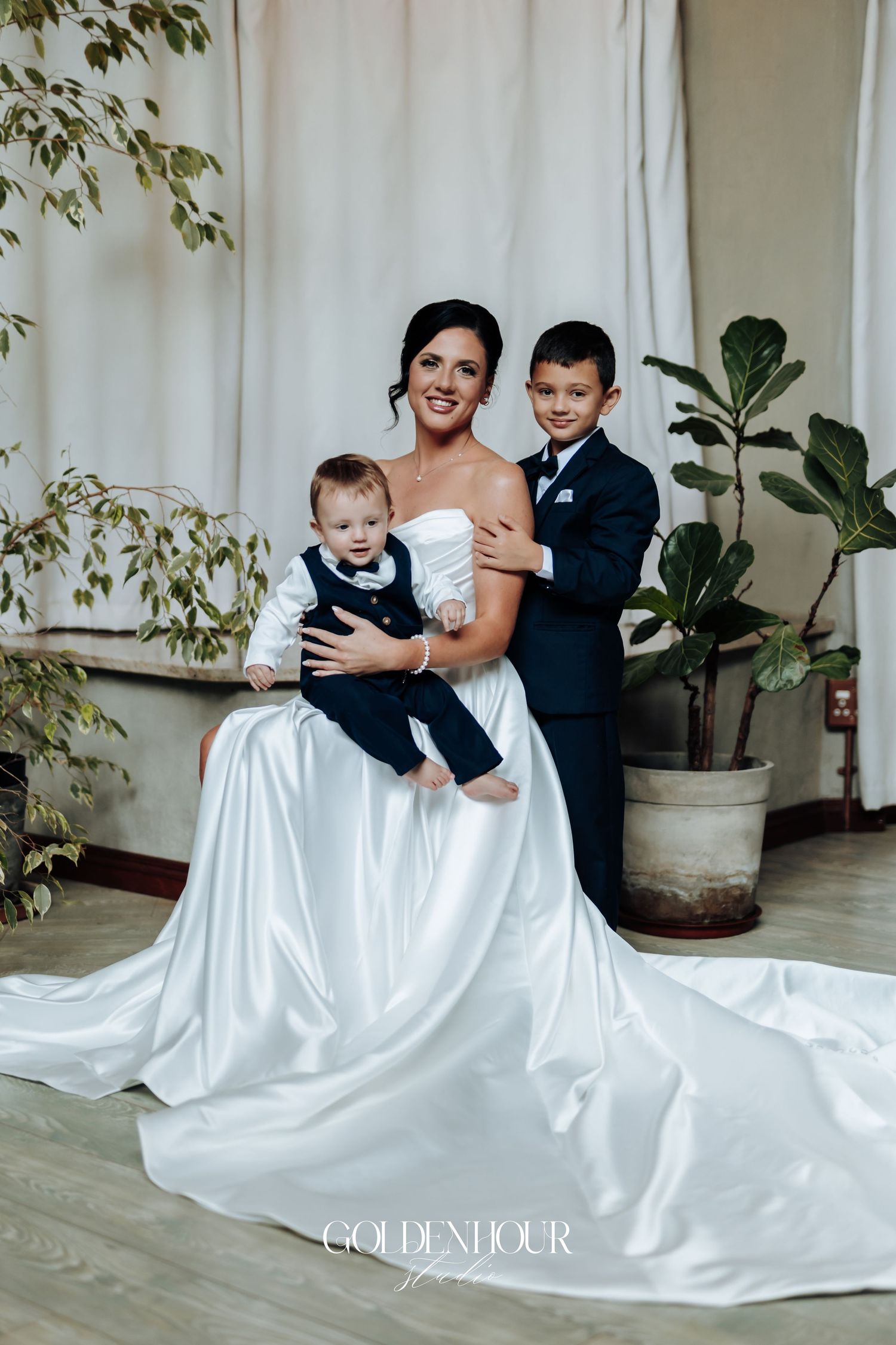 Radiant bride in white strapless gown holding two young boys in navy suits at her wedding.