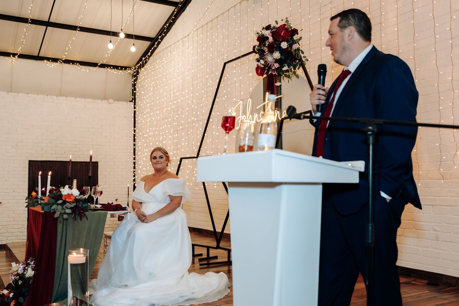 Bride in white gown sits as groom speaks at podium during elegant wedding ceremony with floral decor.