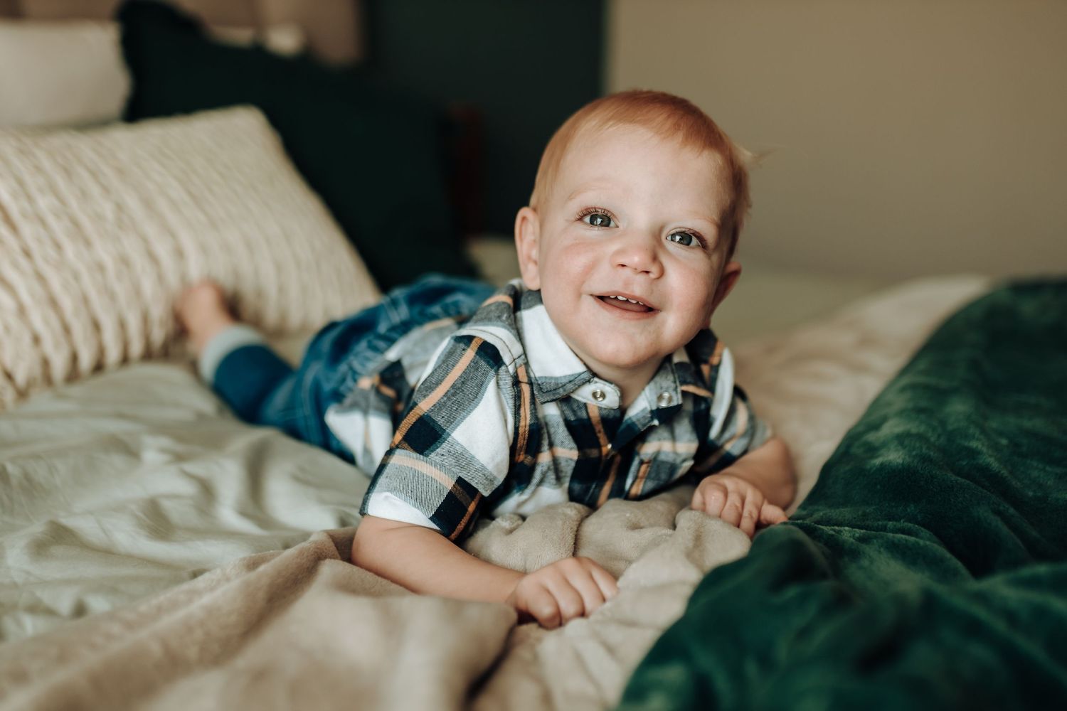 A young child wearing a plaid shirt smiles while lying on bed with green and white bedding.