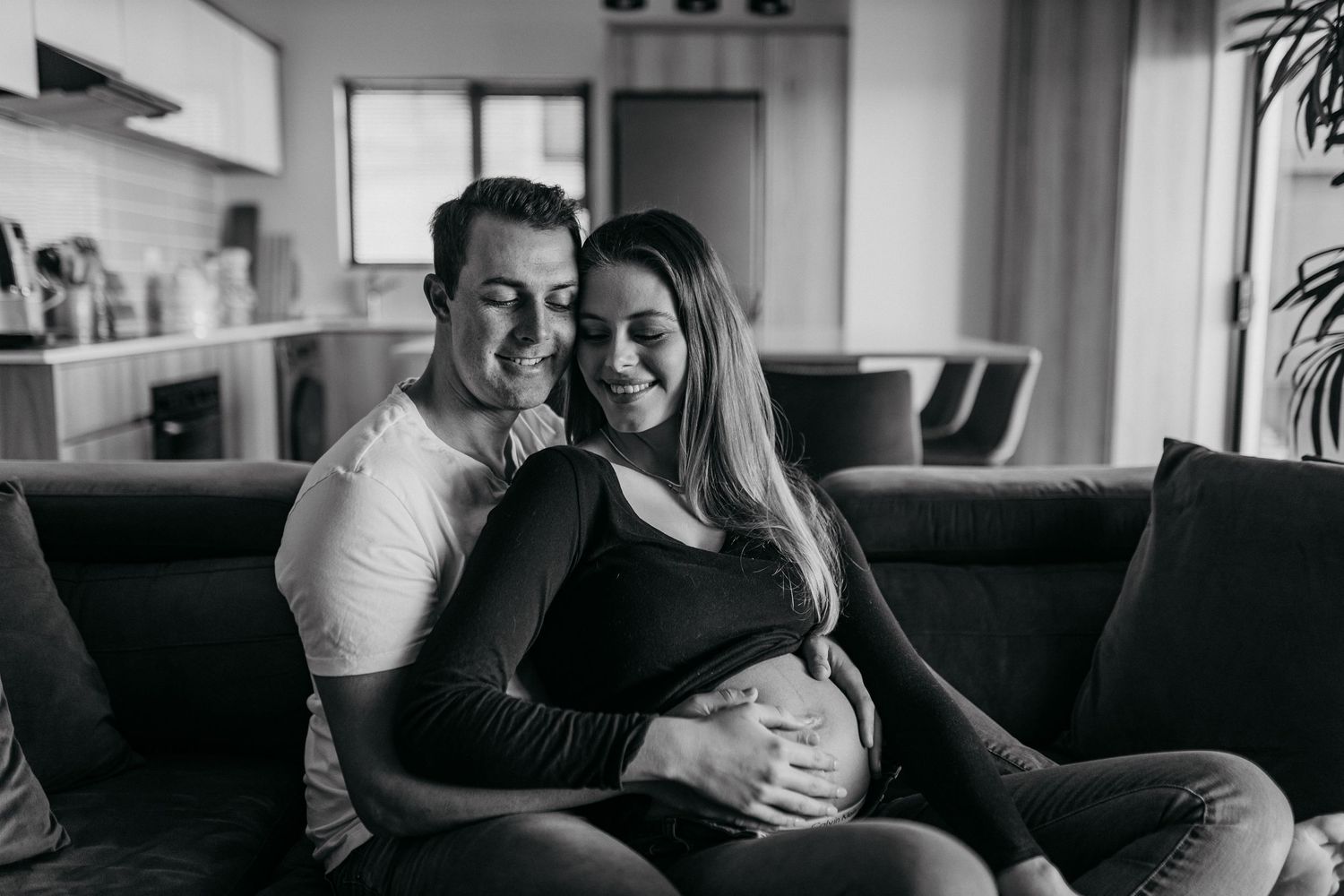 Intimate maternity photoshoot shows couple embracing on dark couch in natural window light.