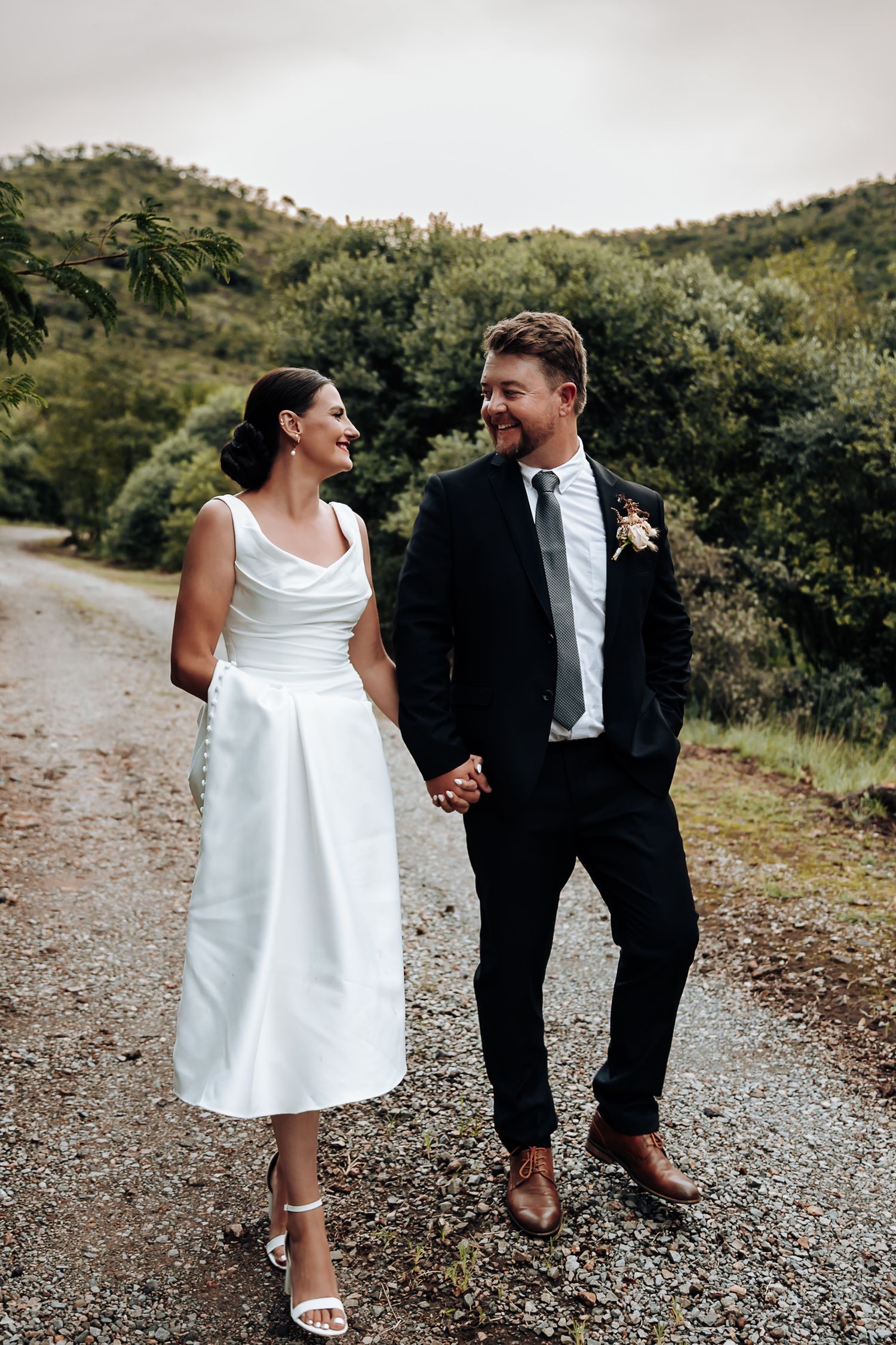 A couple in wedding attire laughs together while walking on a gravel path surrounded by lush greenery.