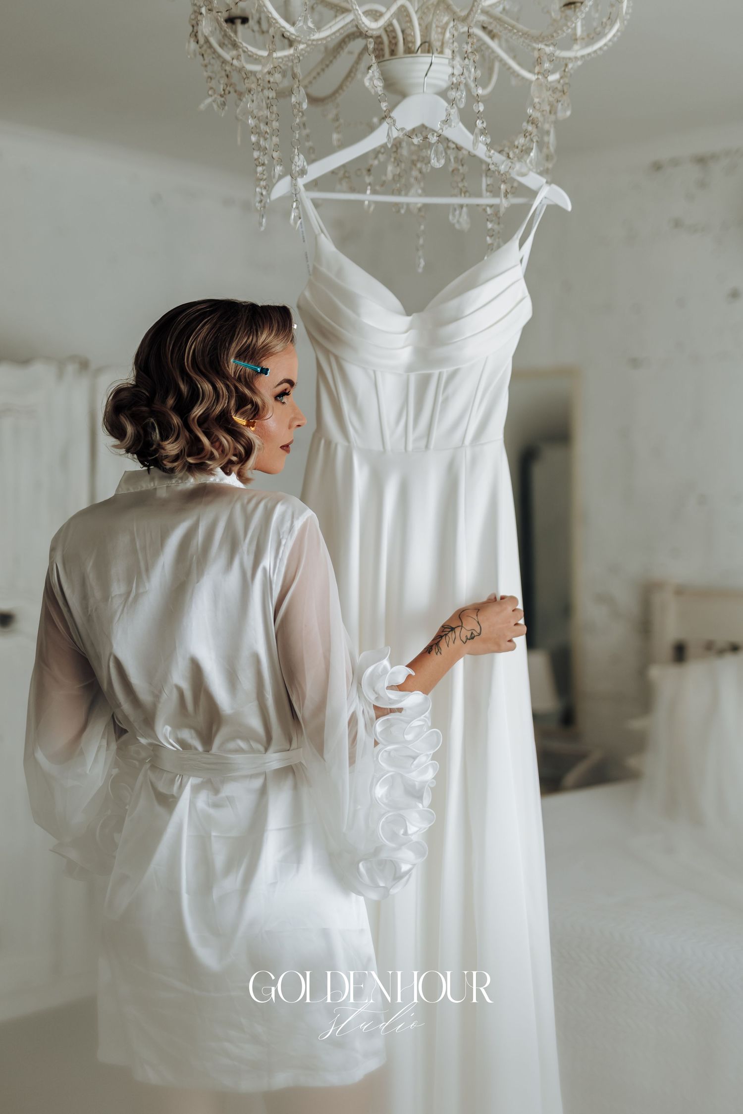 Elegant white wedding dress hanging from crystal chandelier with bride in silk robe preparing for the big day.