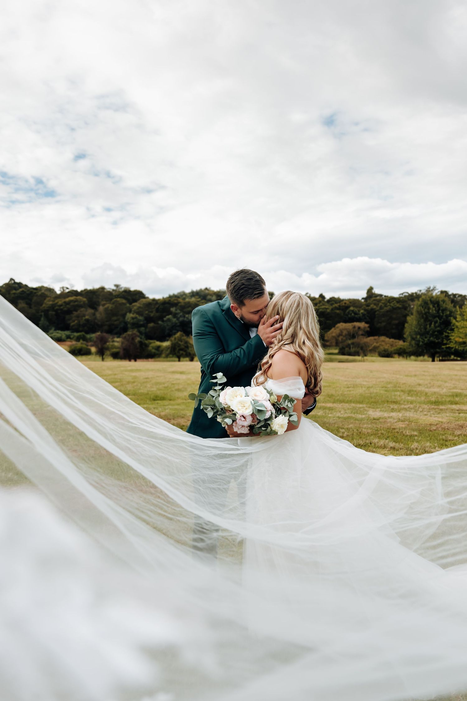A couple shares a romantic kiss outdoors while a flowing white veil creates an ethereal effect in the sunlight.