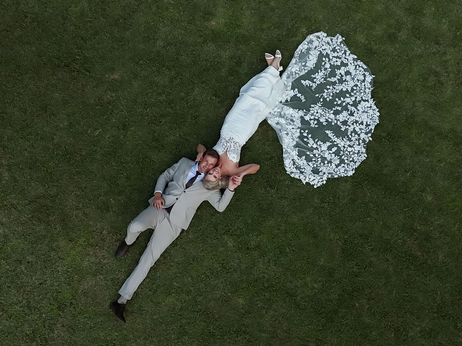 Aerial drone view of newlywed couple lying on green grass, bride in white lace gown with flowing veil, groom in gray suit.