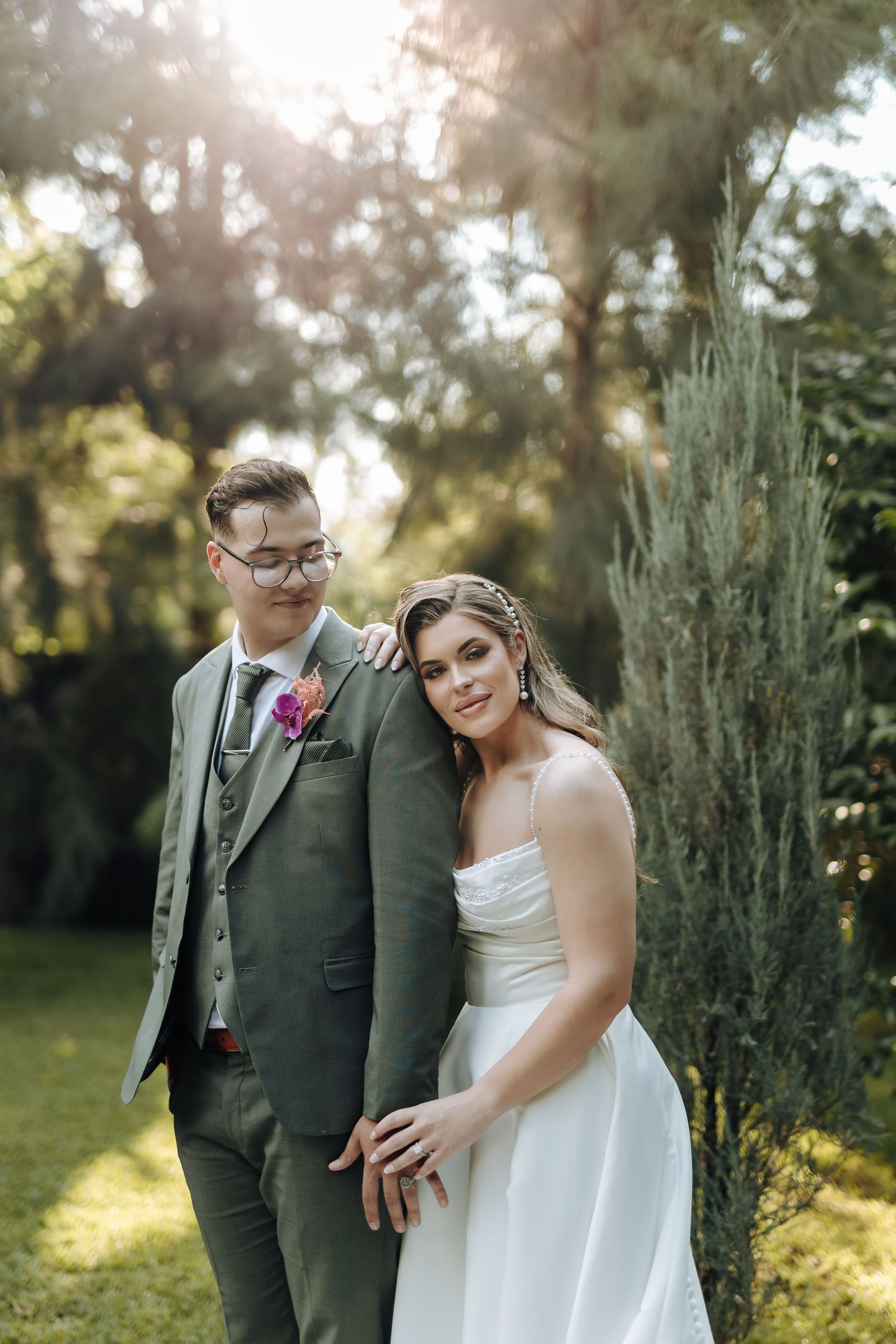 Bride in white gown leans on groom in green suit, posing together in a sunlit garden setting.
