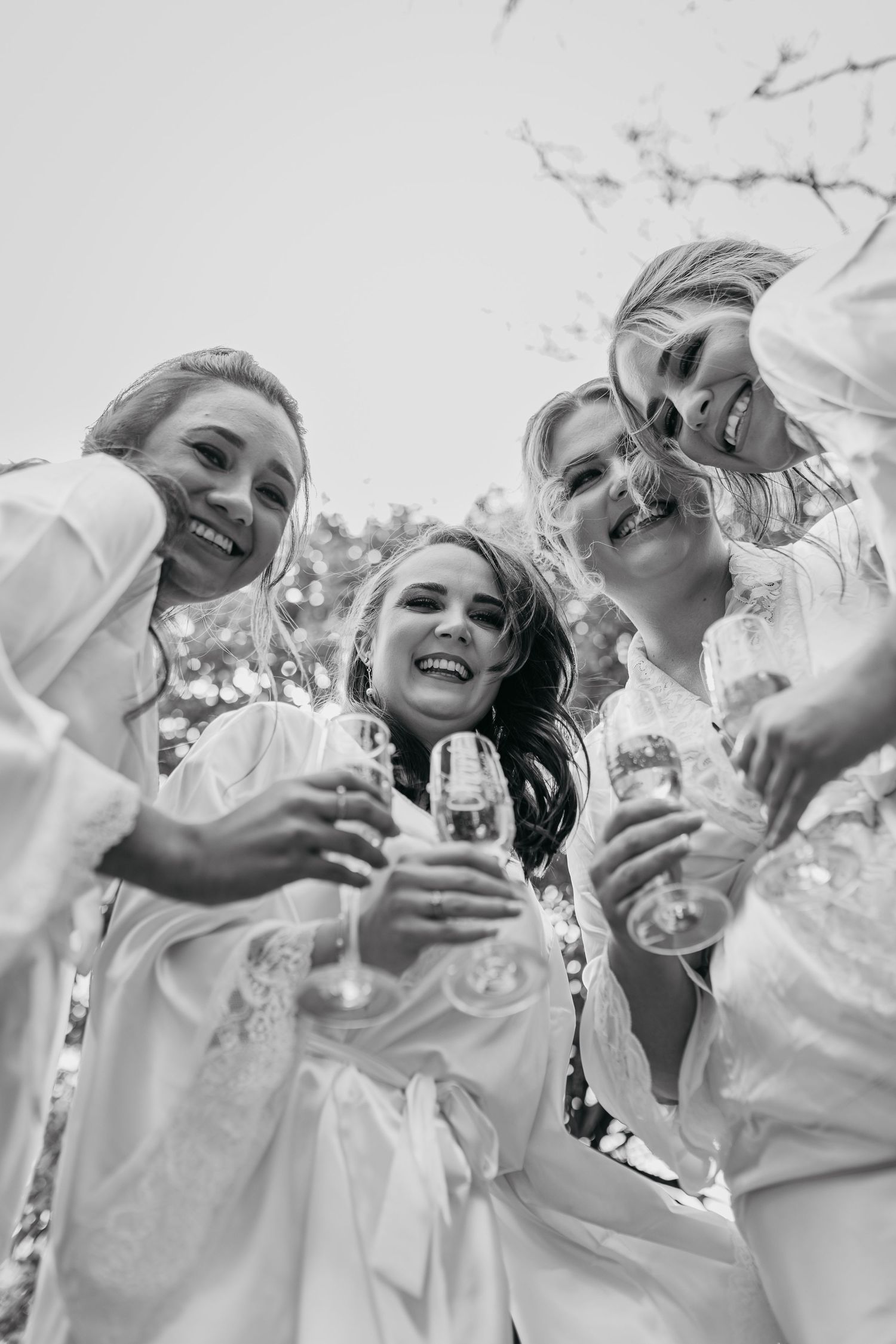 Black and white photo of group wearing white laughing together and holding wine glasses at outdoor celebration.