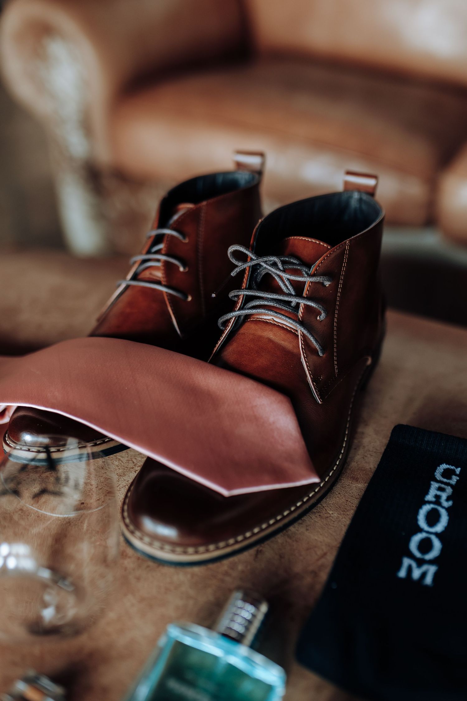 A pair of stylish brown leather chukka boots with white laces sits alongside black packaging and fragrance.