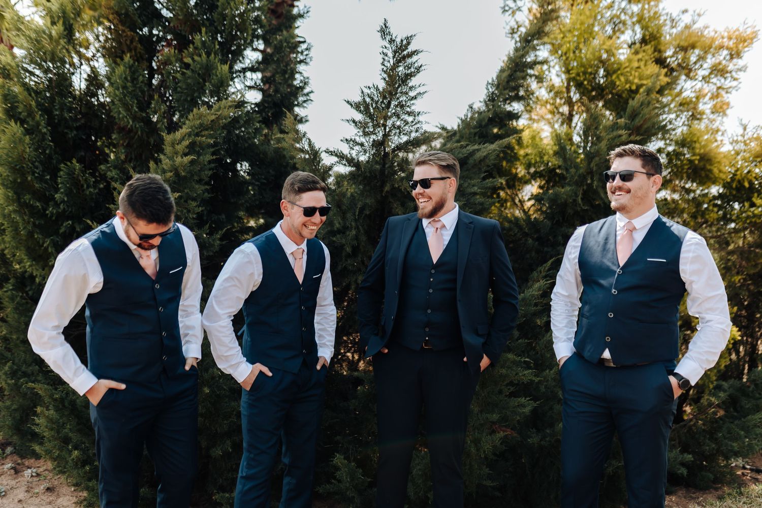 Four people in matching navy waistcoats and dress shirts stand together outdoors in front of evergreen trees.