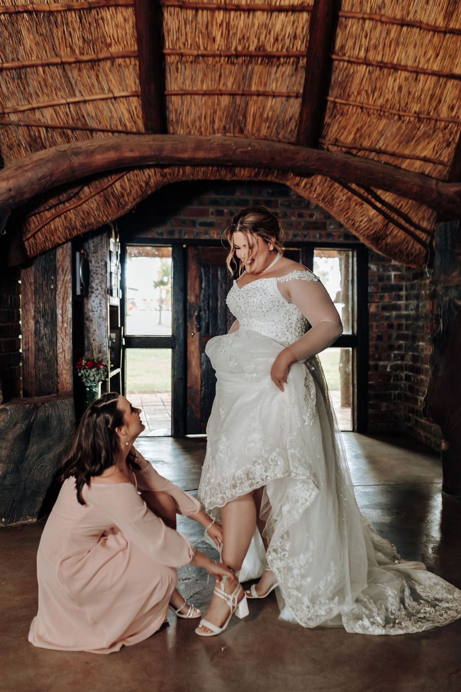 People in a rustic venue with thatched roof ceiling, one helping adjust shoe and dress details in preparation for event.
