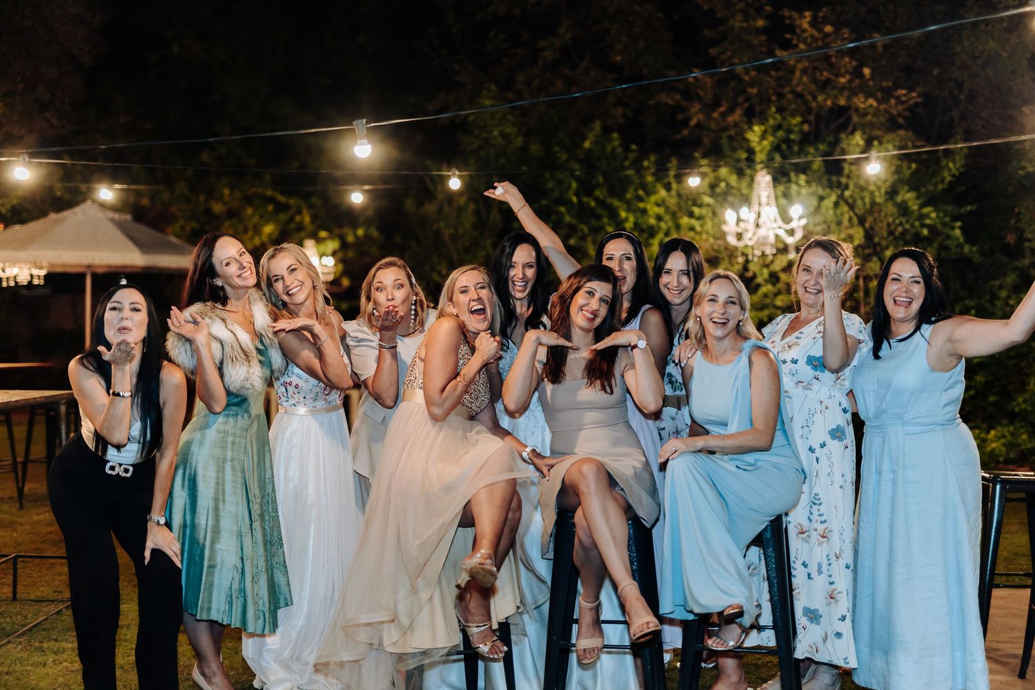 Bride and bridesmaids celebrating at outdoor wedding reception under string lights at night.