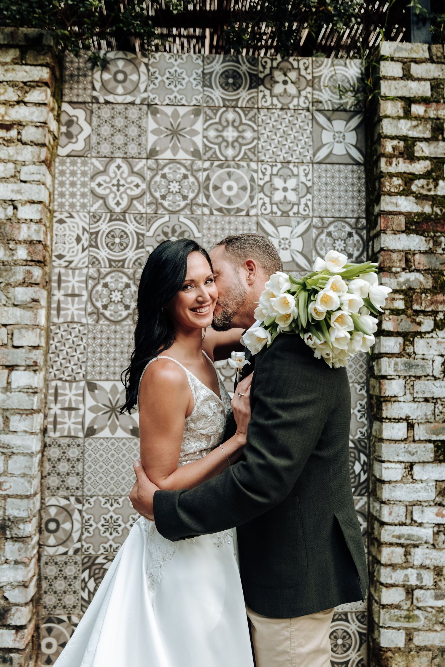 Bride and groom share a romantic moment in front of a decorative tiled wall, bride holding white tulip bouquet.