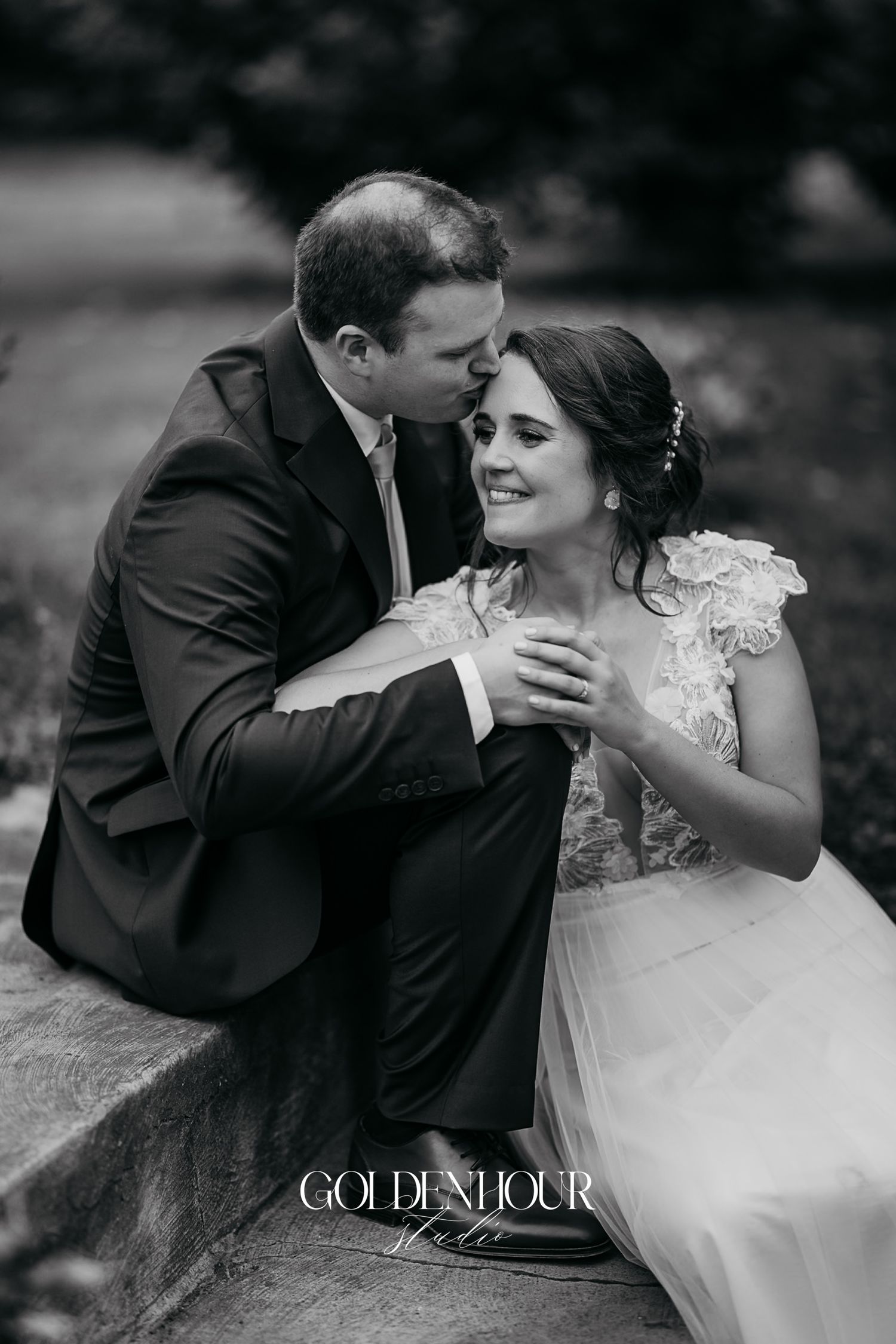Black and white portrait of groom tenderly embracing seated bride outdoors.