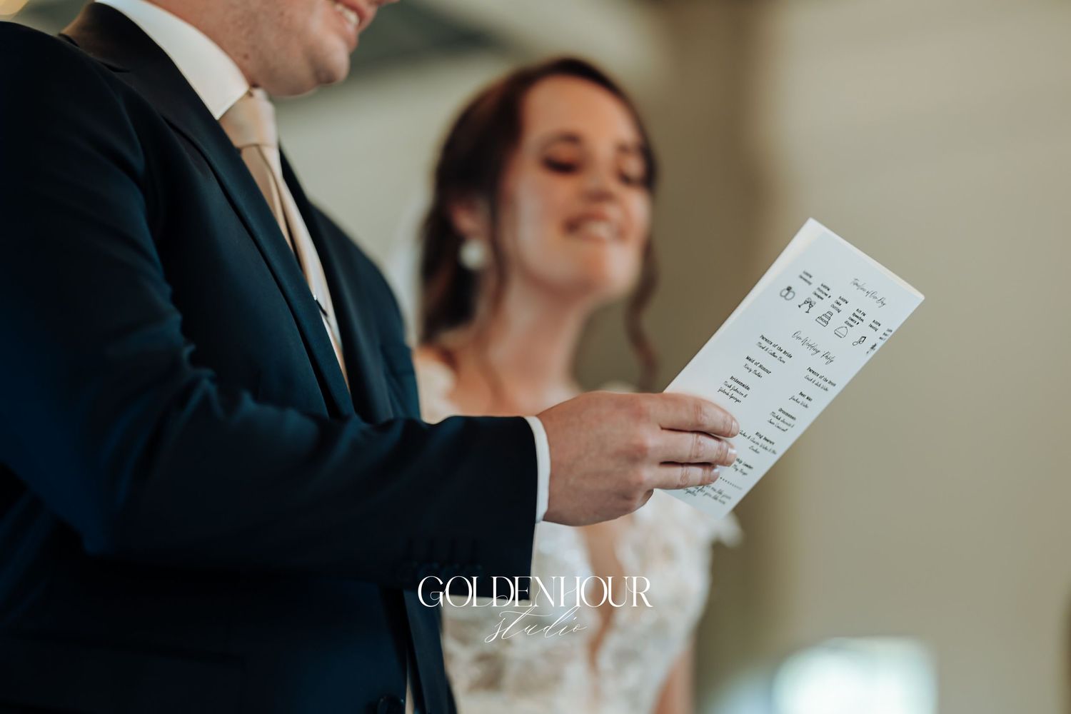 Groom in dark suit reading wedding program while bride smiles beside him.