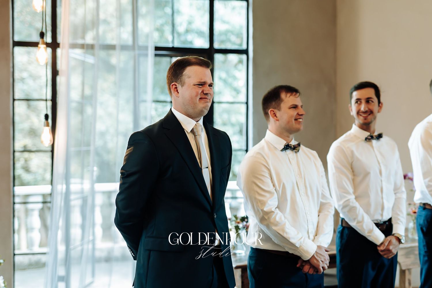 Groom in navy suit stands with groomsmen in white shirts at a bright wedding ceremony venue.