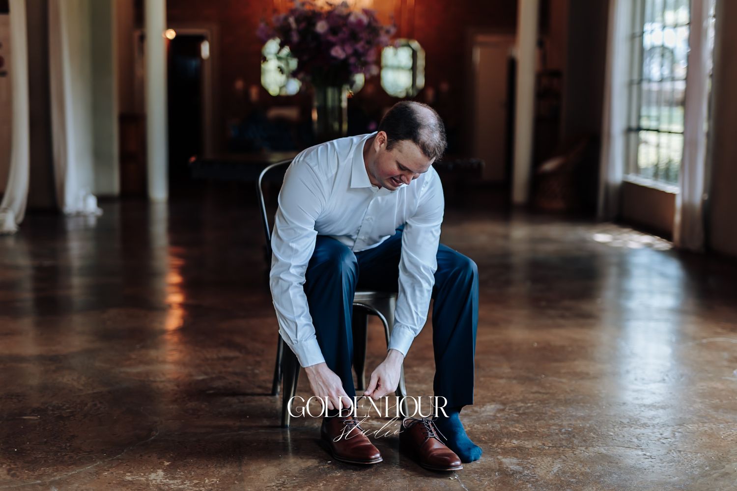 Groom in white shirt and navy pants tying brown dress shoes before wedding.