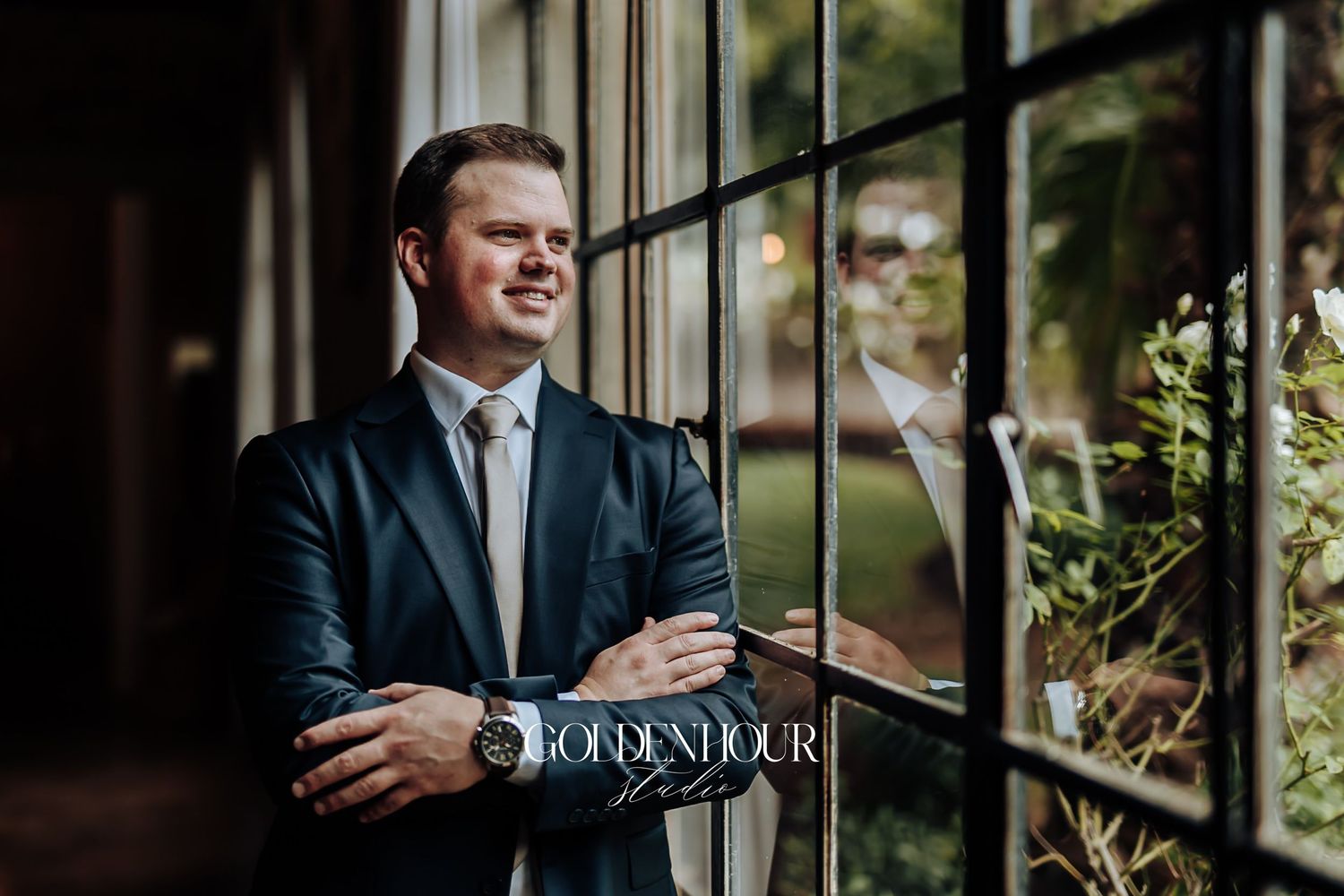 Handsome groom in a dark suit smiling while leaning against large windows with garden views.