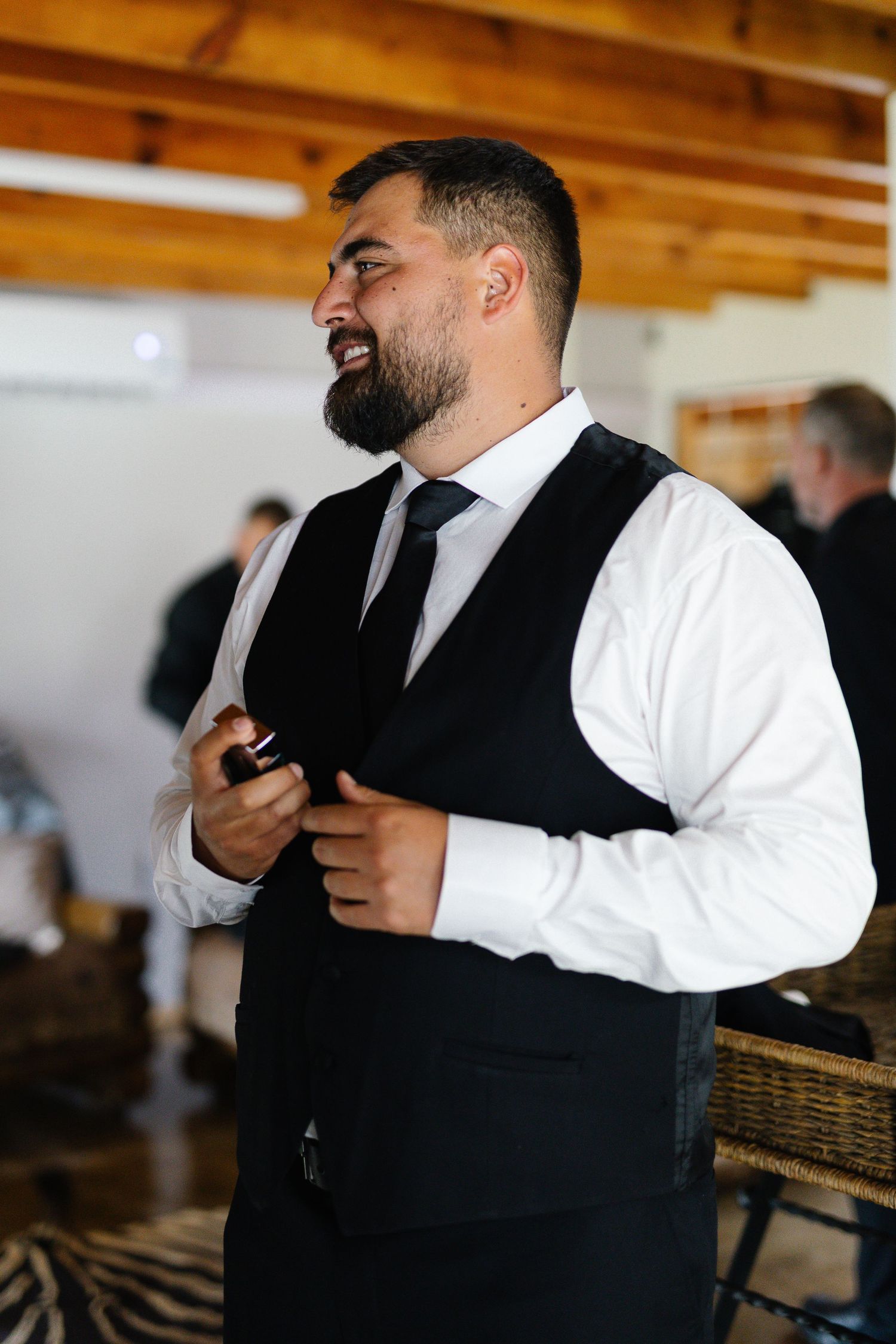 A bearded man in formal attire with a black vest and tie smiles while standing in a room with wooden beams.