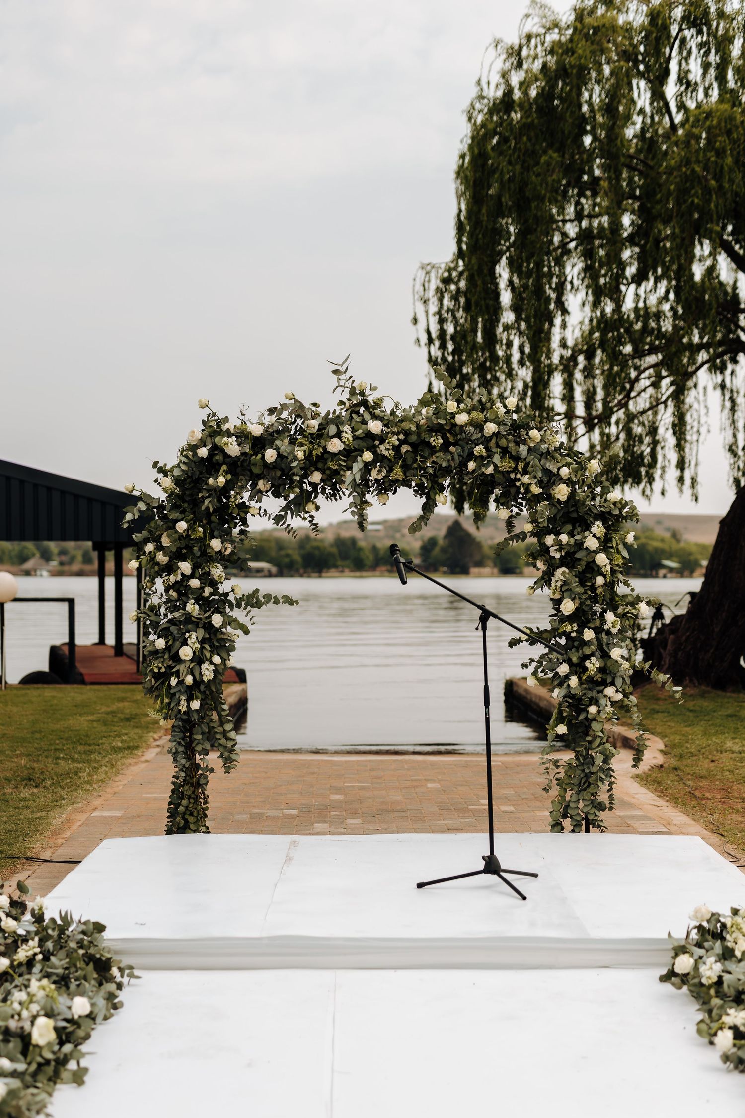 A panoramic view of an outdoor wedding ceremony setup with a white aisle runner and floral arch overlooking a lake.