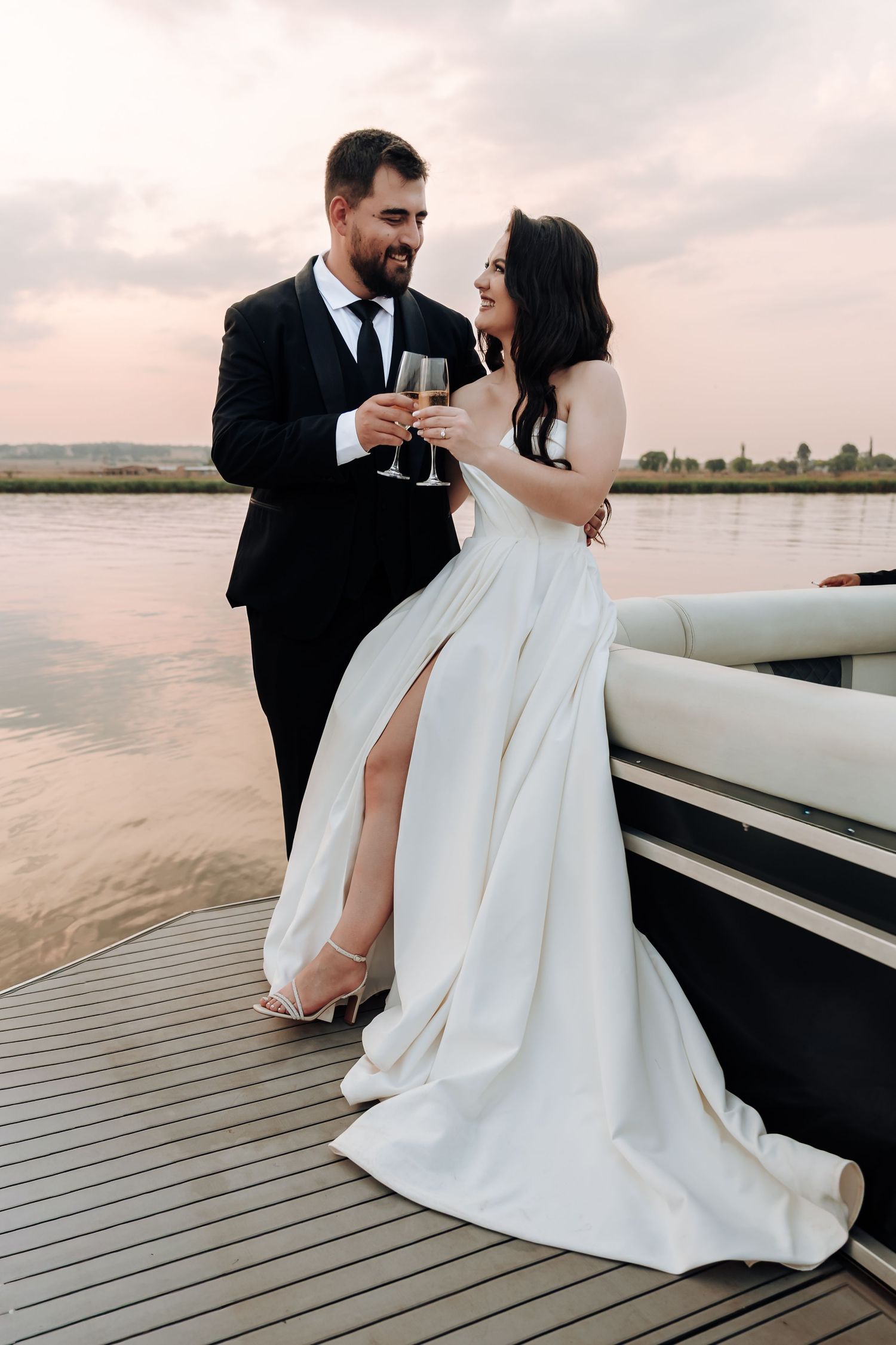 A romantic wedding couple shares champagne on a wooden deck overlooking a serene waterfront at sunset.