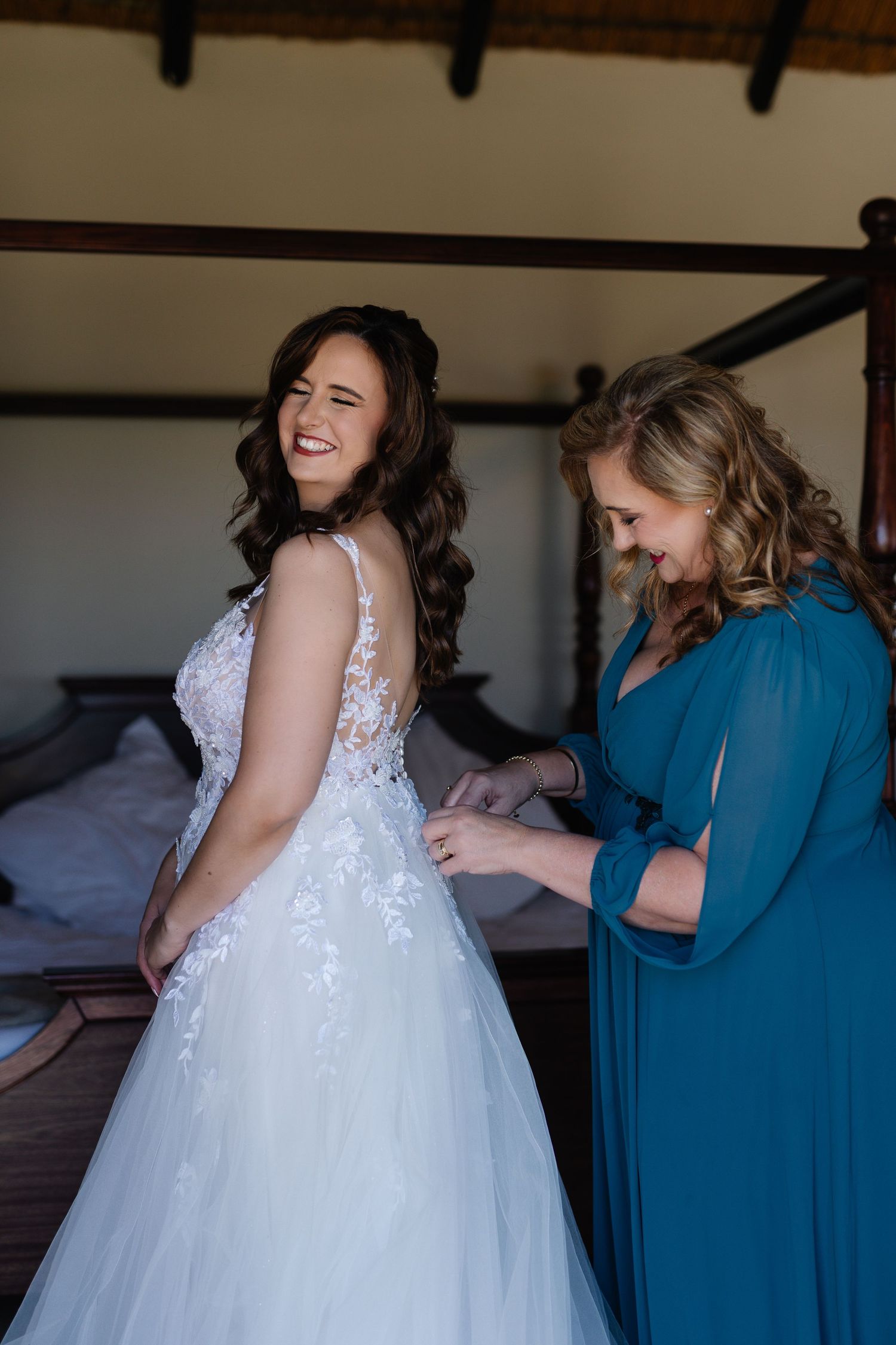 A joyful moment during wedding preparations as someone in a teal dress helps button up a white lace wedding gown.