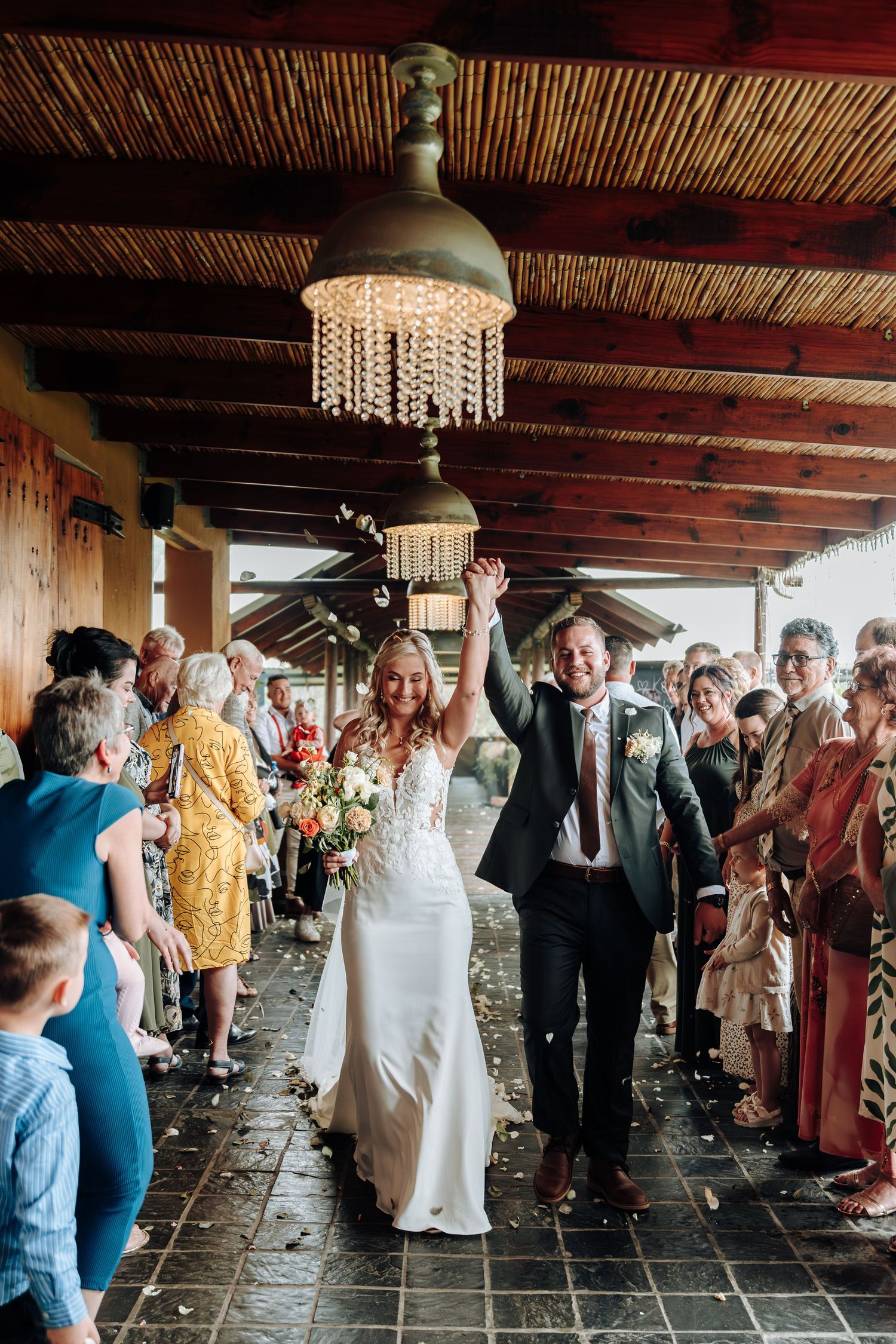 Wedding guests gather under a rustic covered porch with wooden beams and pendant lights during a celebration.