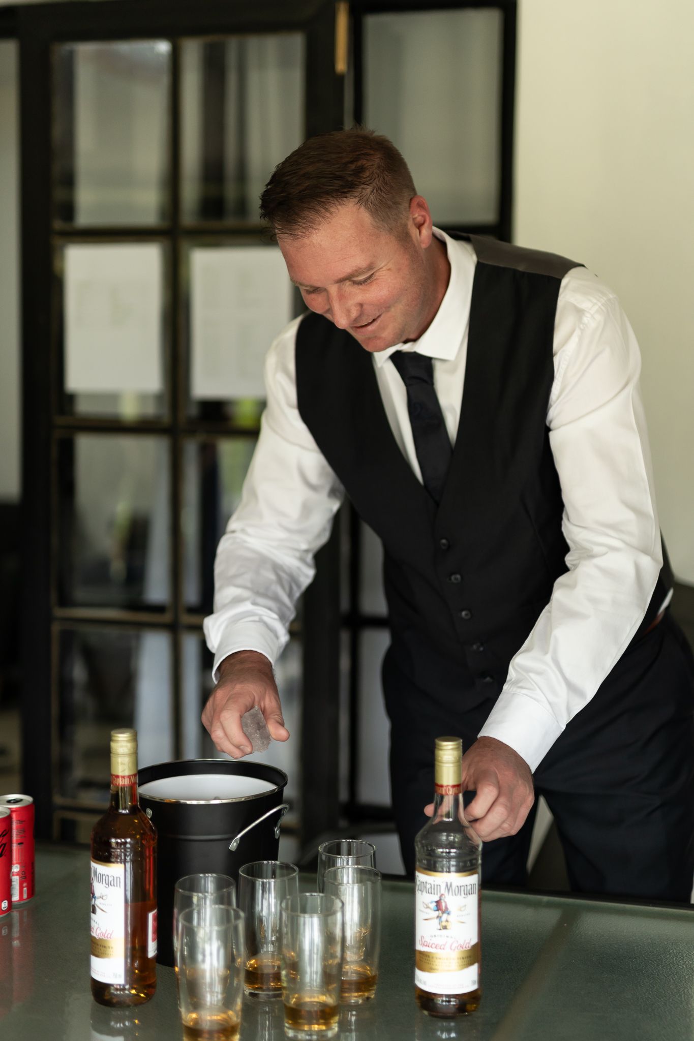 A bartender in formal attire pours drinks at a bar setup with whiskey bottles and glasses.