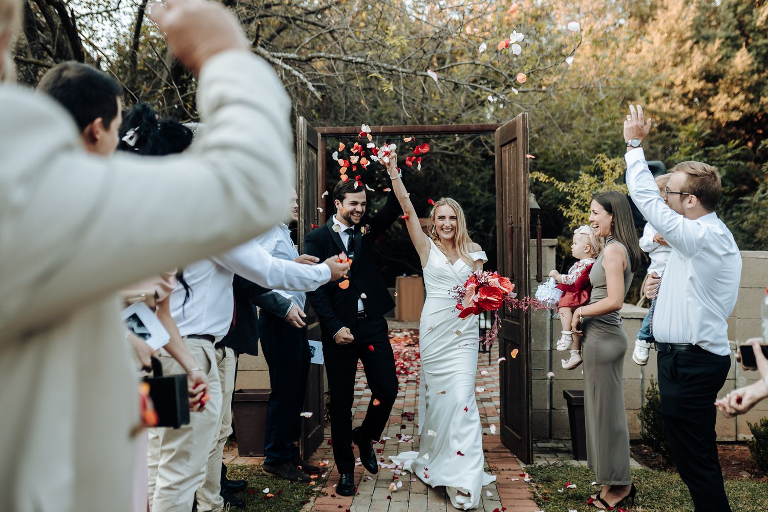 Joyous wedding exit with guests throwing confetti as couple walks through garden archway.