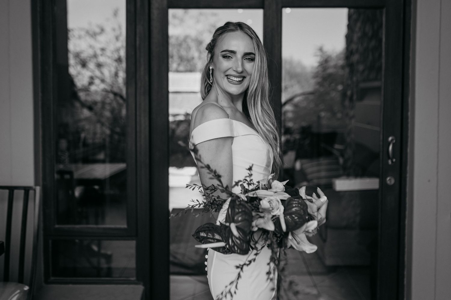 Black and white photograph of smiling bride holding floral bouquet by glass doors.