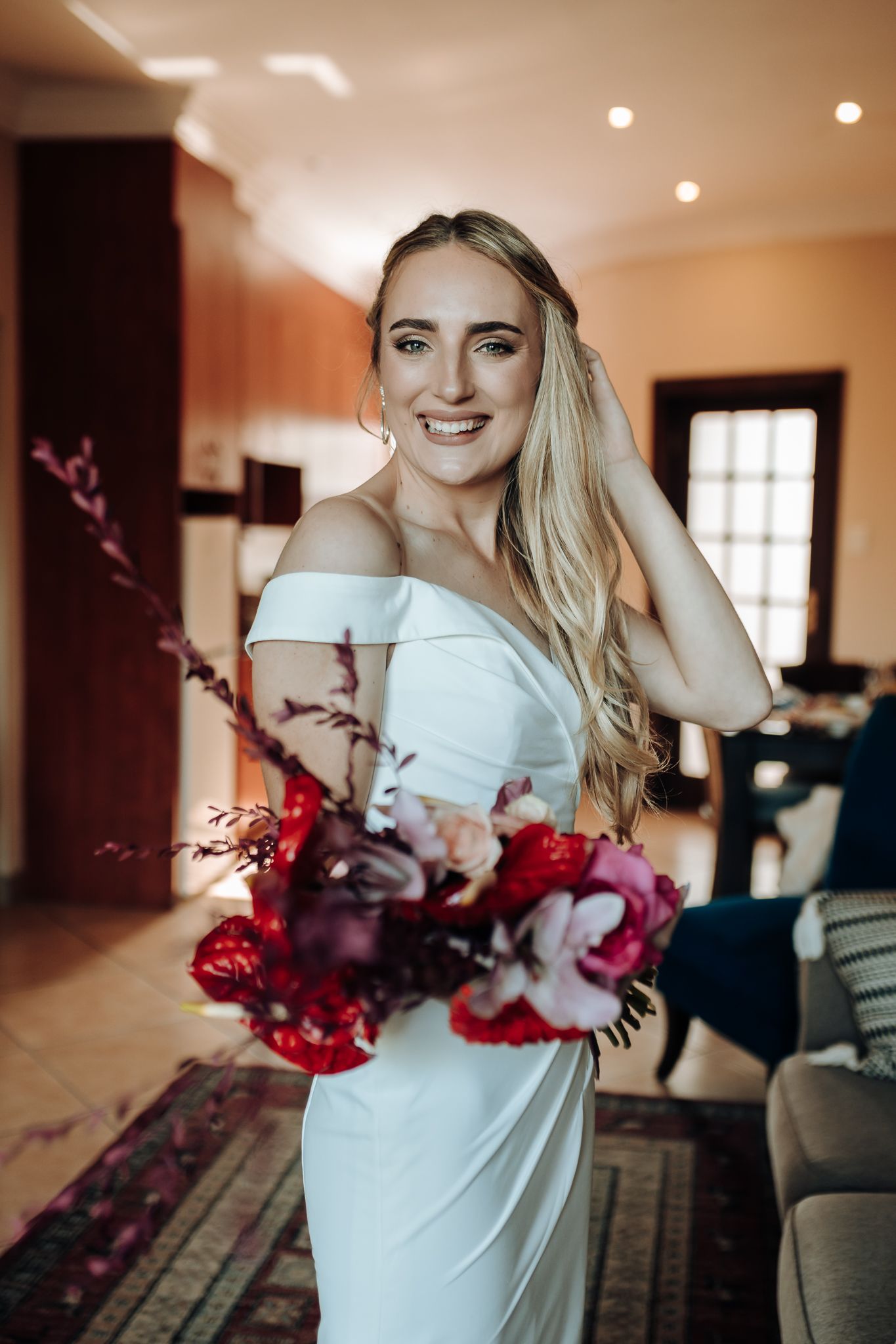 Bride in an off-shoulder white dress holding vibrant red and pink flowers.