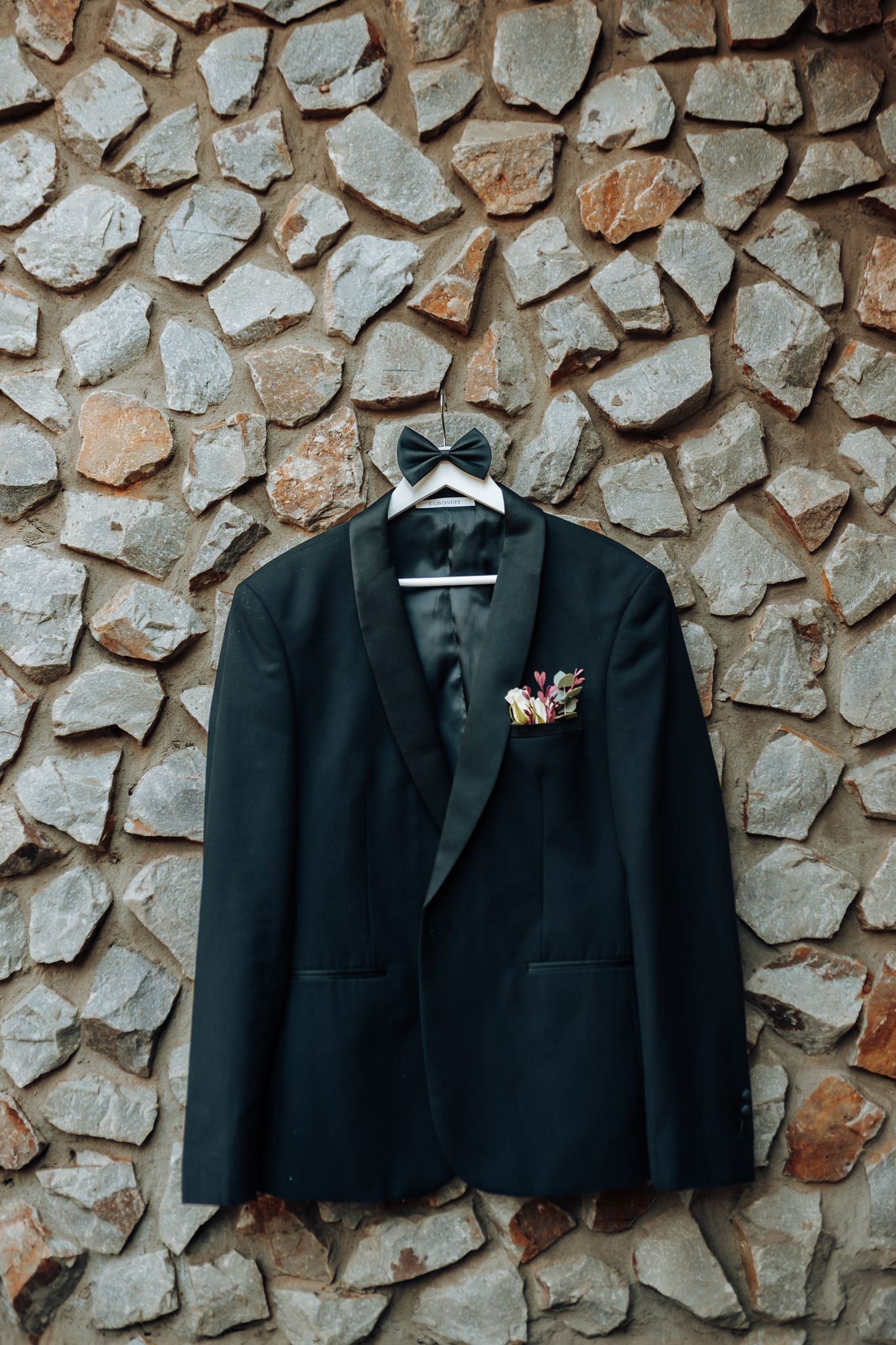 Black tuxedo jacket with bowtie and boutonniere hanging on stone wall.